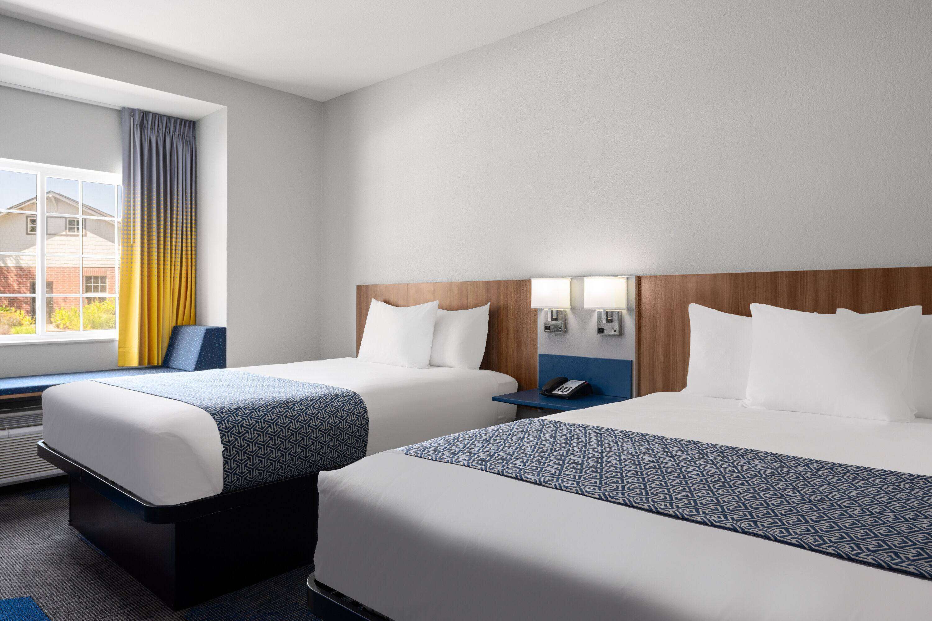 A hotel room with two beds, a window with curtains, a bedside table with a phone, and a view of buildings outside.