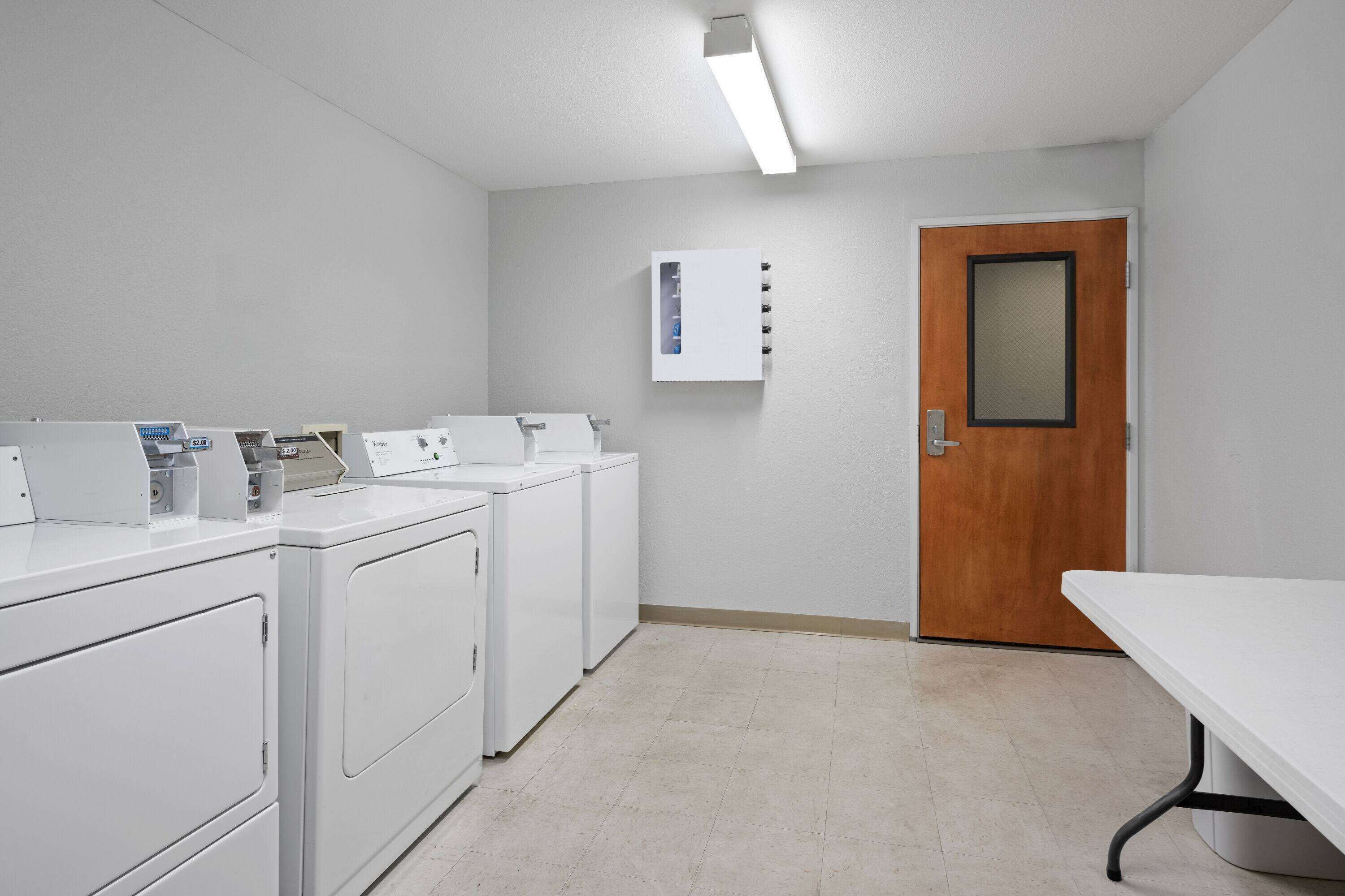 A laundry room with four white washers and dryers, a wooden door, and a white table.