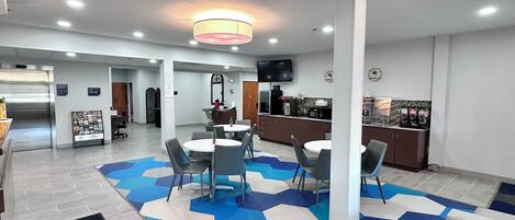 A modern hotel lobby with a reception desk, a dining area with round tables and chairs, a kitchen area, and a patterned floor.