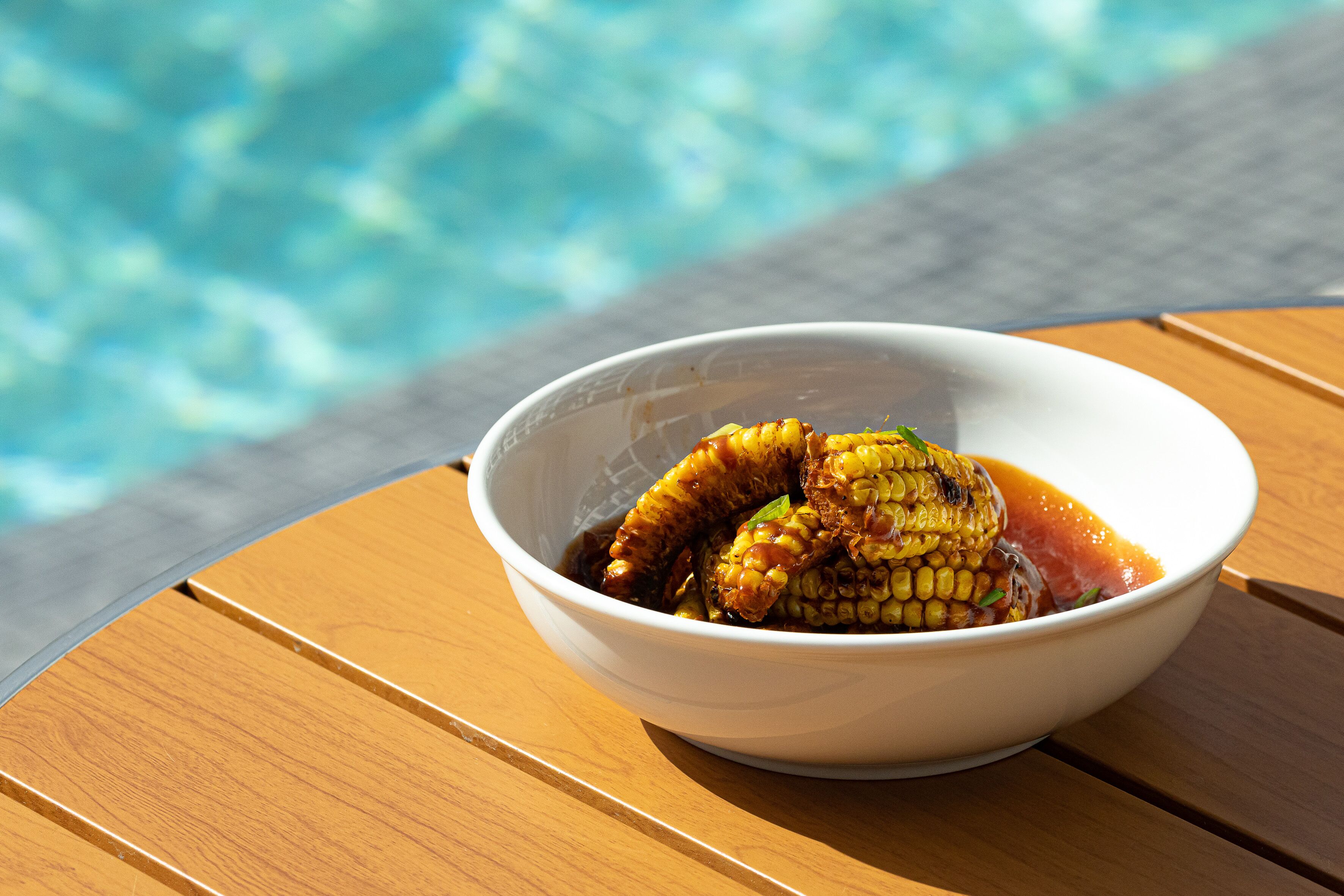 Un plato de elotes con salsa, sobre una mesa de madera cerca de una piscina.
