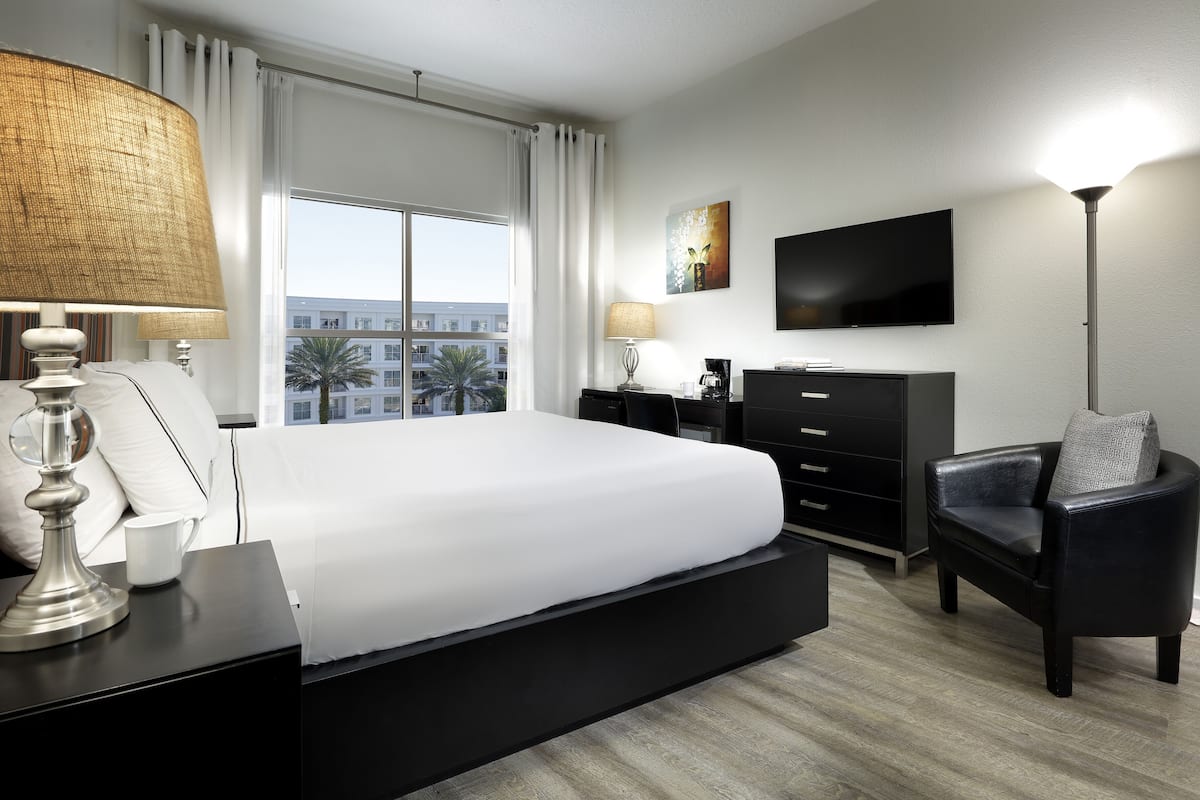 A hotel room with a large bed, a desk with a lamp, a chair, a TV, and a view of buildings outside the window.