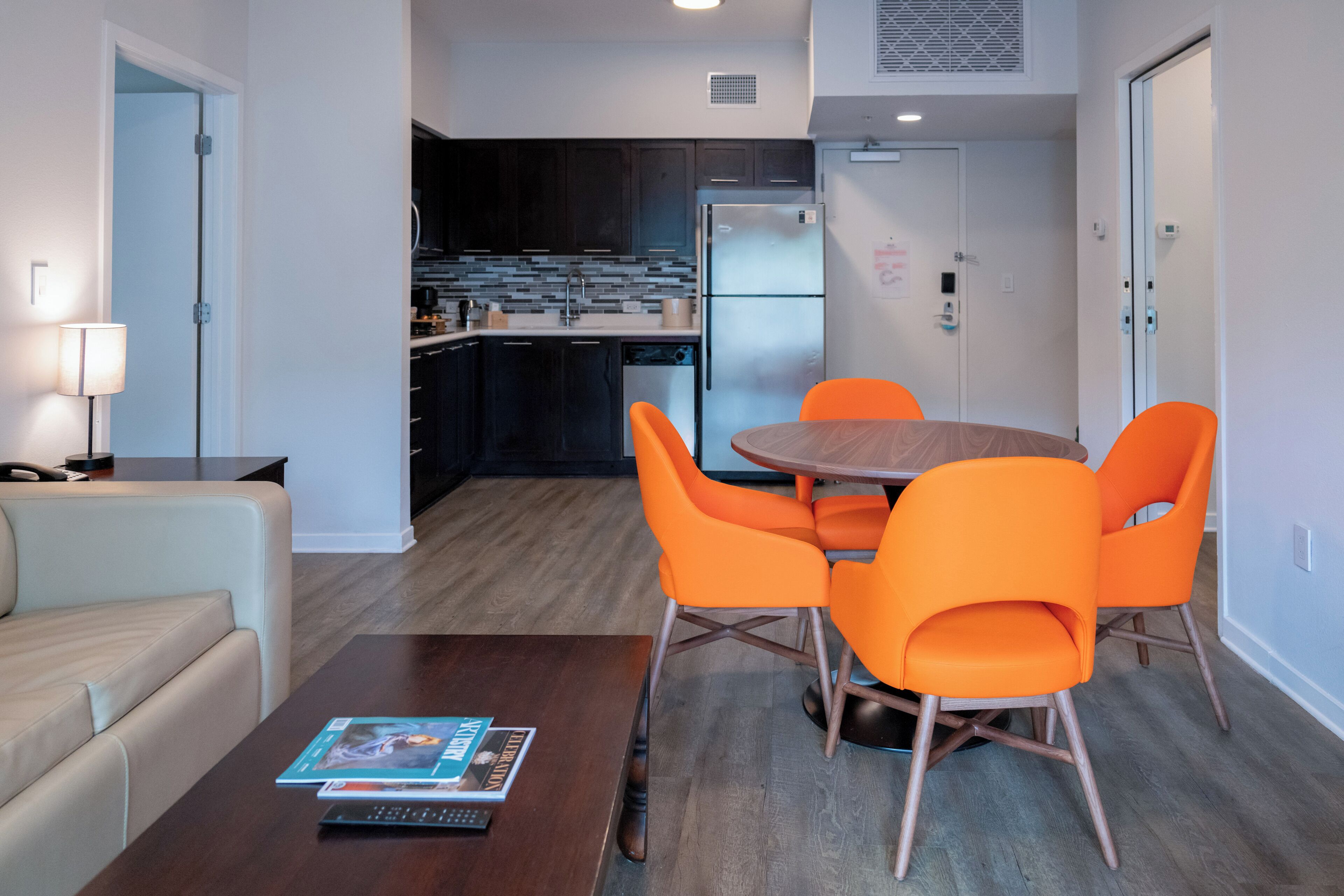 Eine moderne Küche mit dunklen Schränken, einem Edelstahlkühlschrank und einem Essbereich mit einem runden Holztisch und orangefarbenen Stühlen.