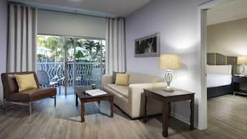 Een hotelkamer met een bank, fauteuil, salontafel en een klein tafeltje met een lamp. Er is een balkon met uitzicht op palmbomen en een gebouw.