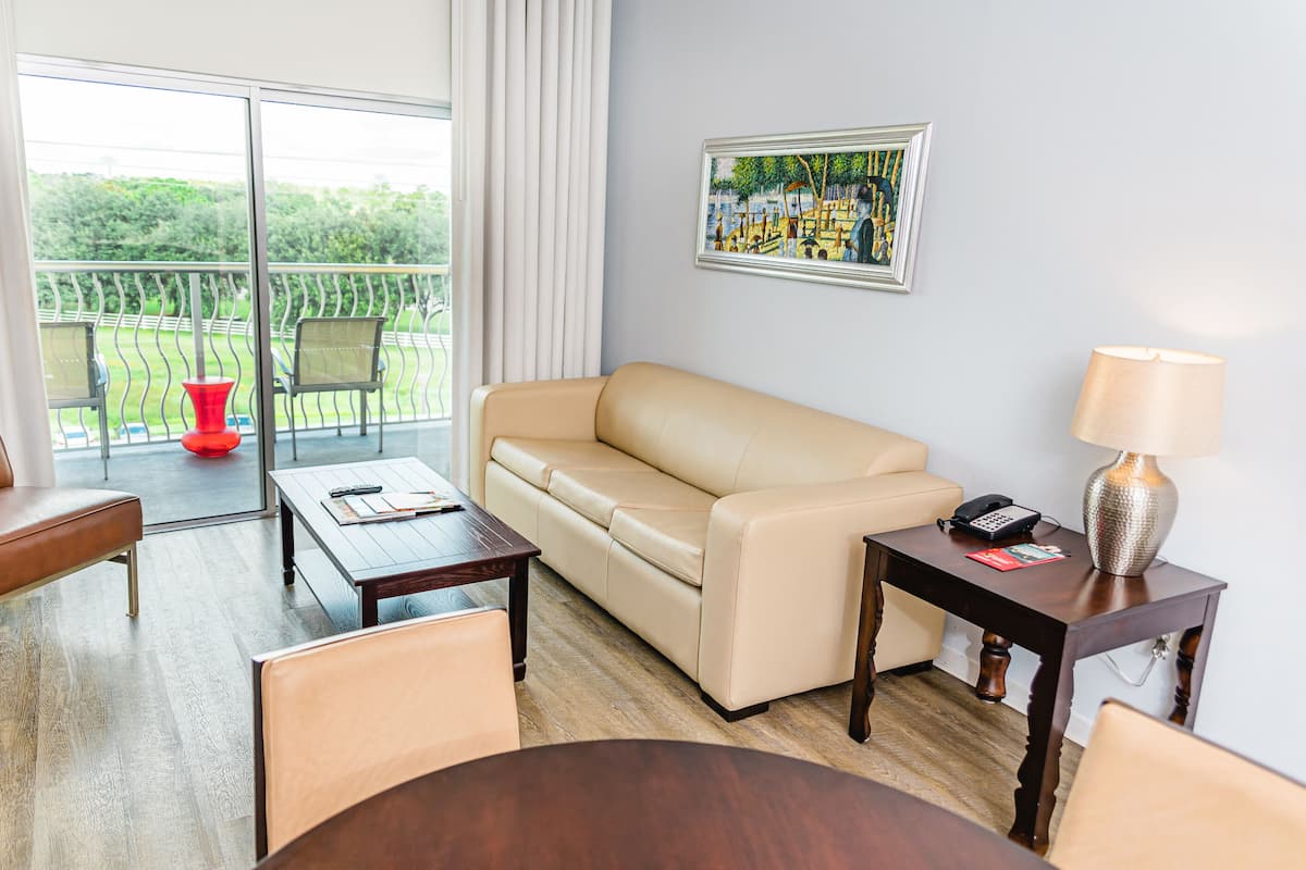 A hotel room with a beige sofa, a wooden coffee table, a desk with a lamp, and a painting on the wall.