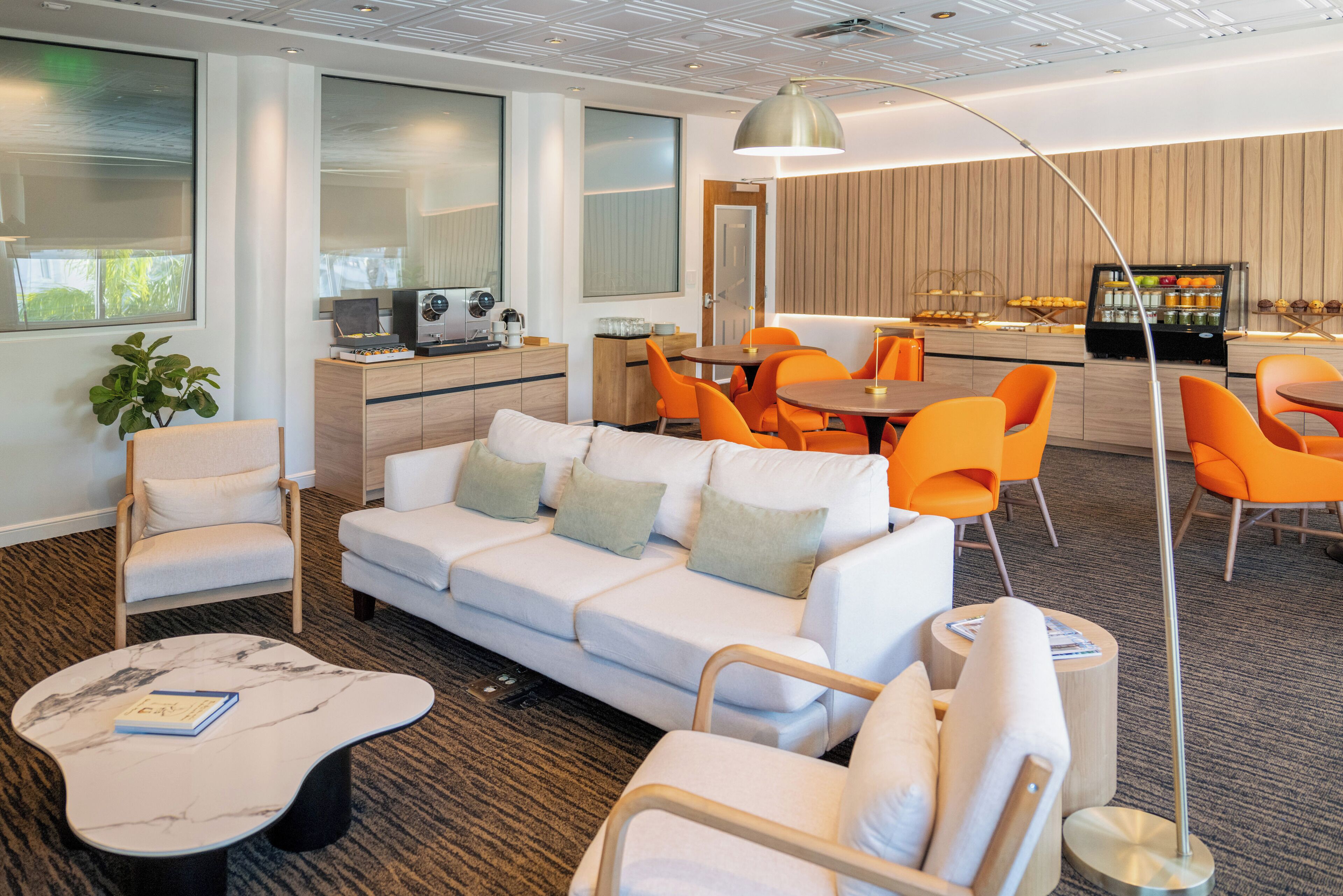 A modern hotel lobby with a white sofa, orange chairs, a marble coffee table, and a bar area with a refrigerator.