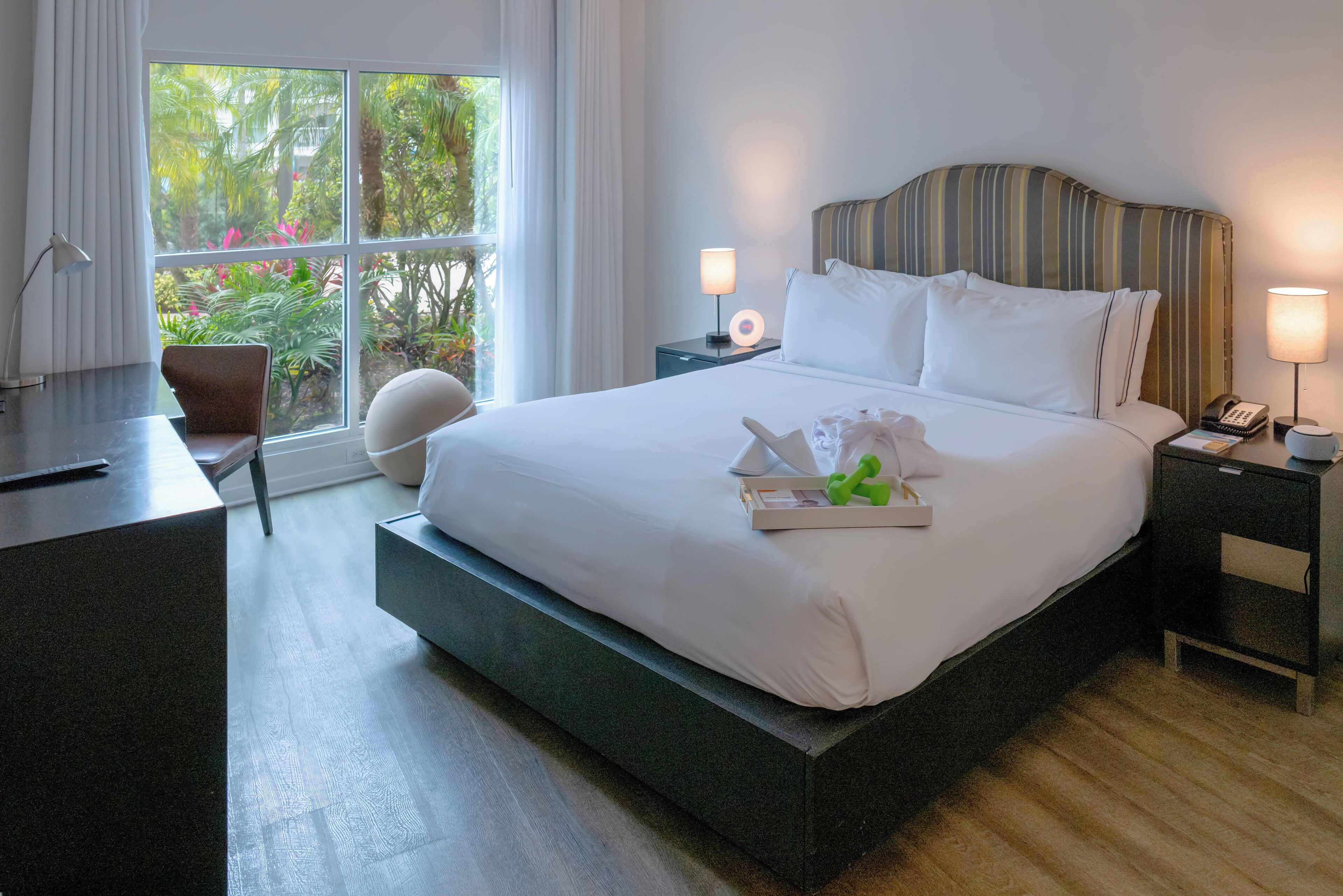 A hotel room with a large bed, a desk, a chair, and a window with a view of greenery.