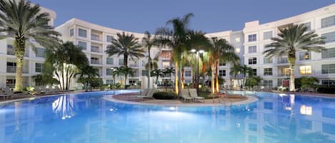 A hotel pool area with a clear blue pool, palm trees, and lounge chairs.