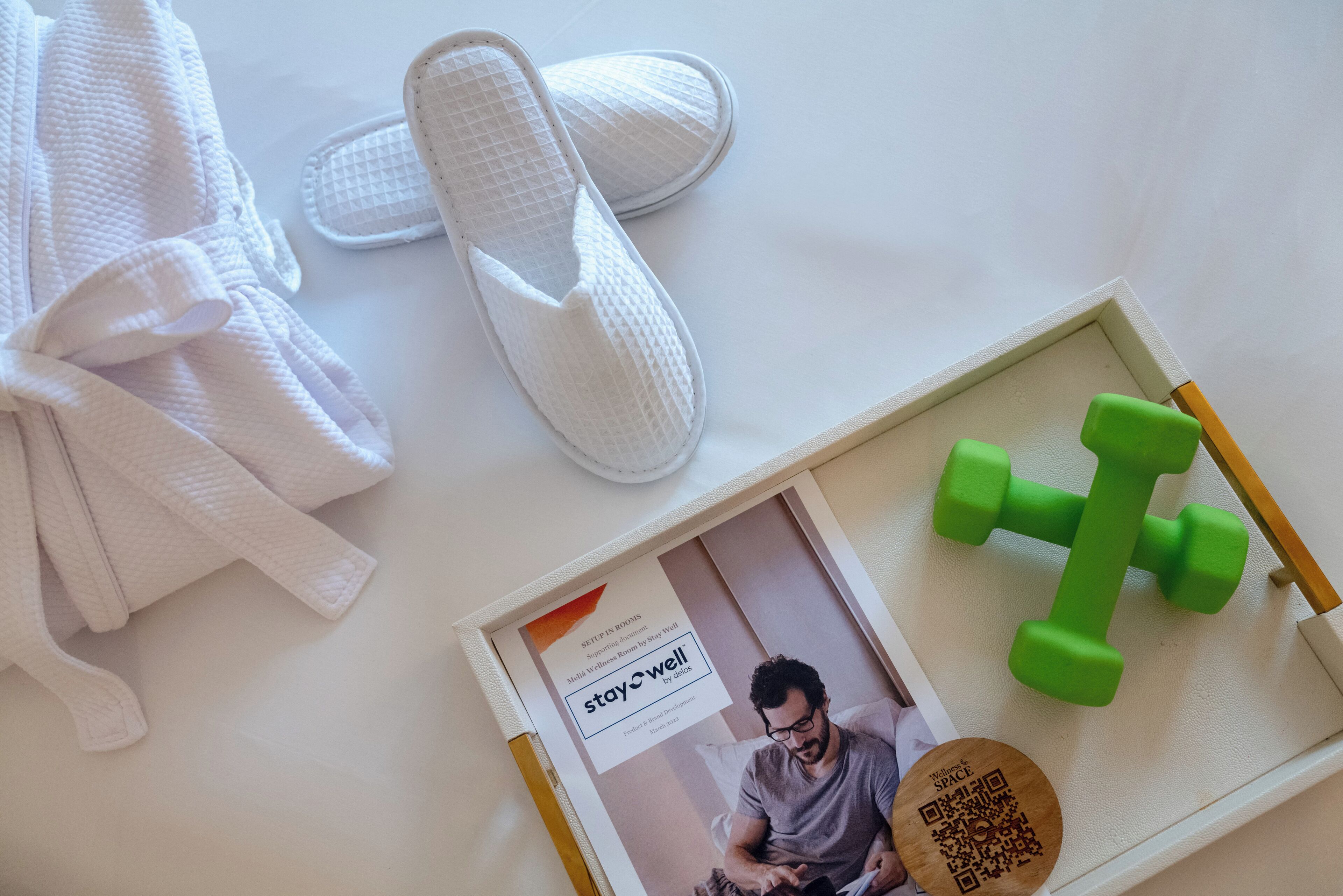 A neatly arranged room with a white bathrobe, white slippers, a magazine, and a green dumbbell on a white surface.