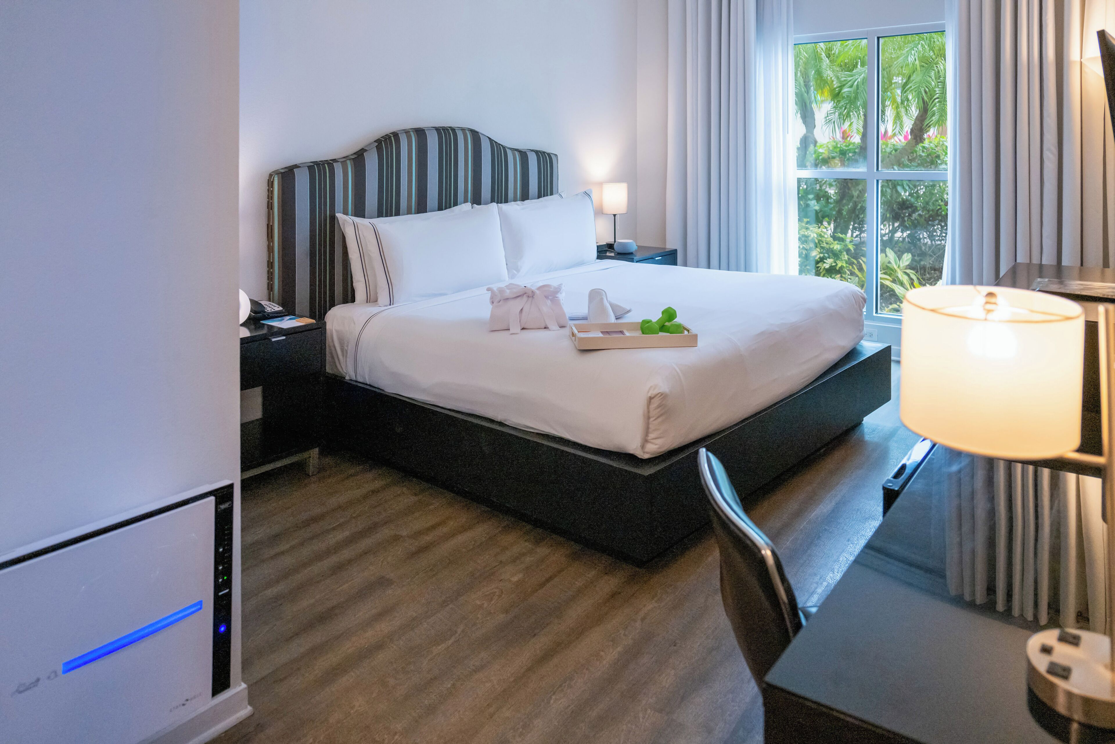 Una habitación de hotel con cama, mesita de noche, lámpara y vistas a zonas verdes por la ventana.