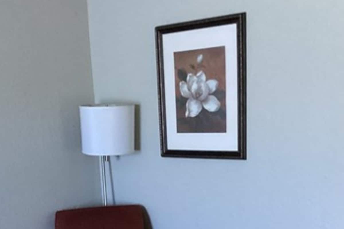 A room with a red armchair, a small dark table, a white floor lamp, and a framed picture of a flower on the wall.