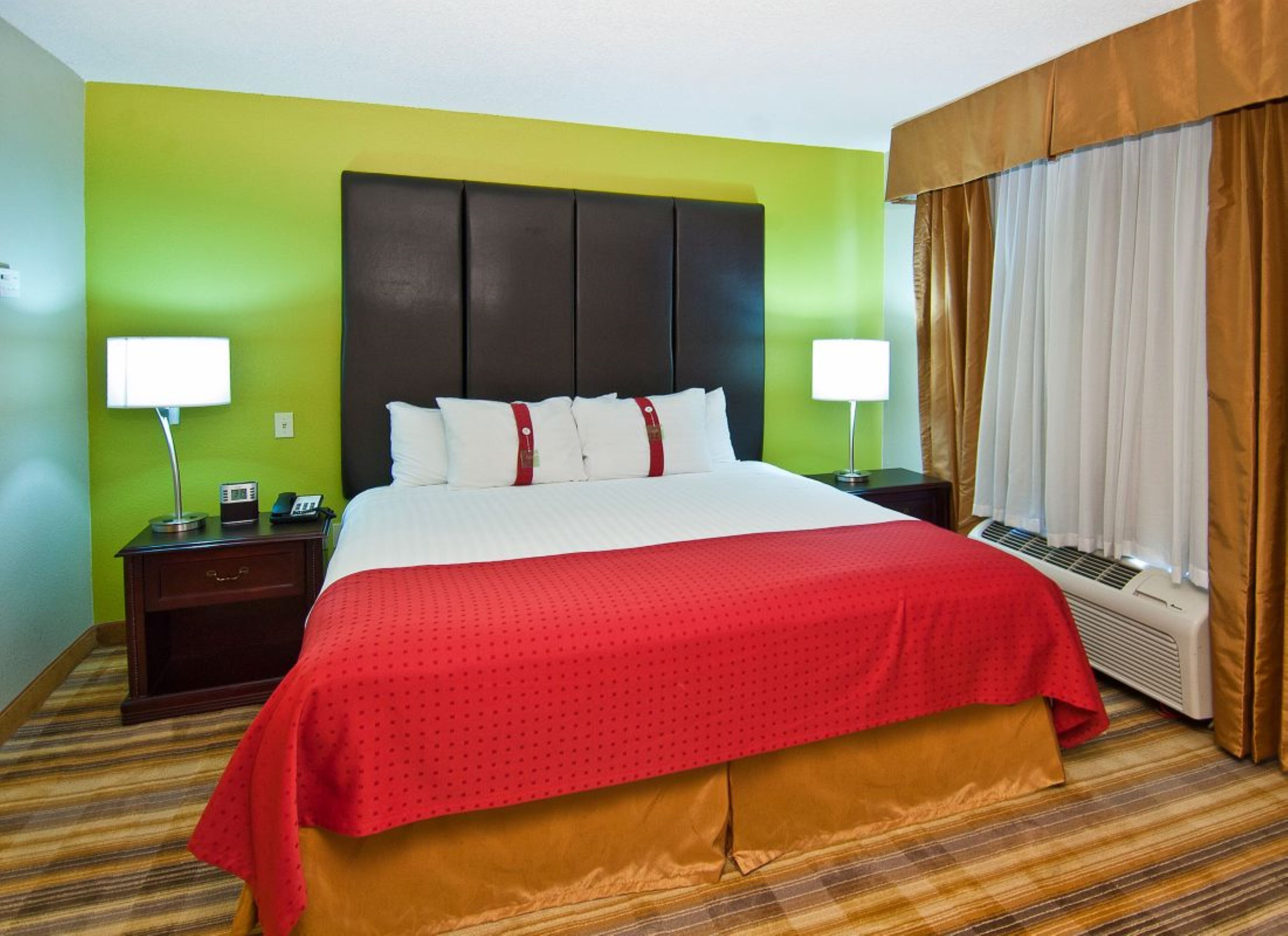 A hotel room with a large bed, two bedside tables with lamps, a window with curtains, and a wall heater.