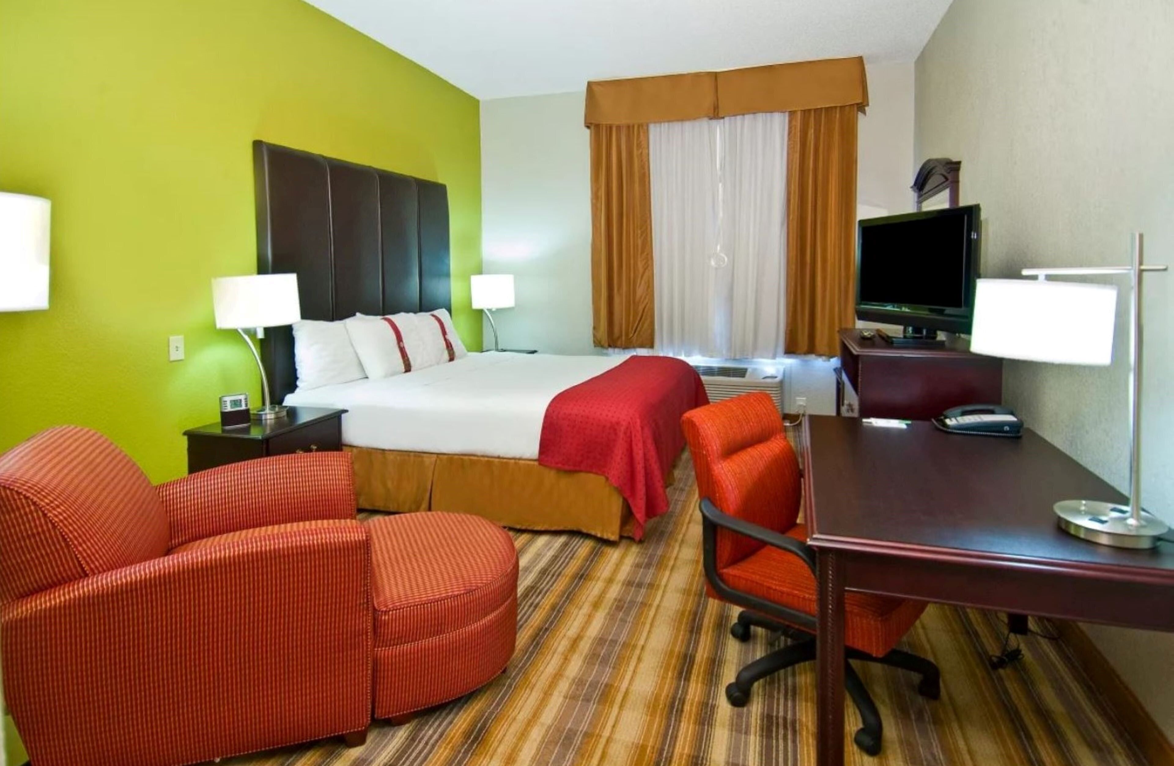 A hotel room with a bed, two chairs, a desk, a TV, and a window with curtains.