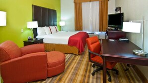 Quarto de hotel com cama, duas cadeiras, escrivaninha, televisão e janela com cortinas.