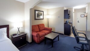 A hotel room with a bed, a red sofa, a small table, a desk, a chair, a lamp, a framed picture on the wall, and a microwave.