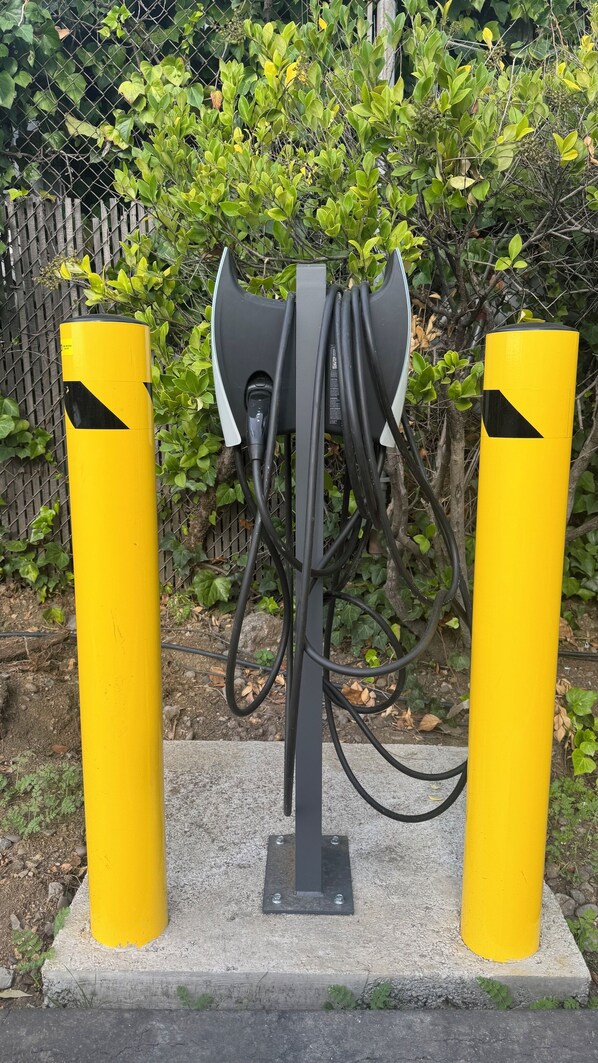 A yellow electric vehicle charging station with cables connected to a central unit, set against a backdrop of greenery and a chain-link fence.