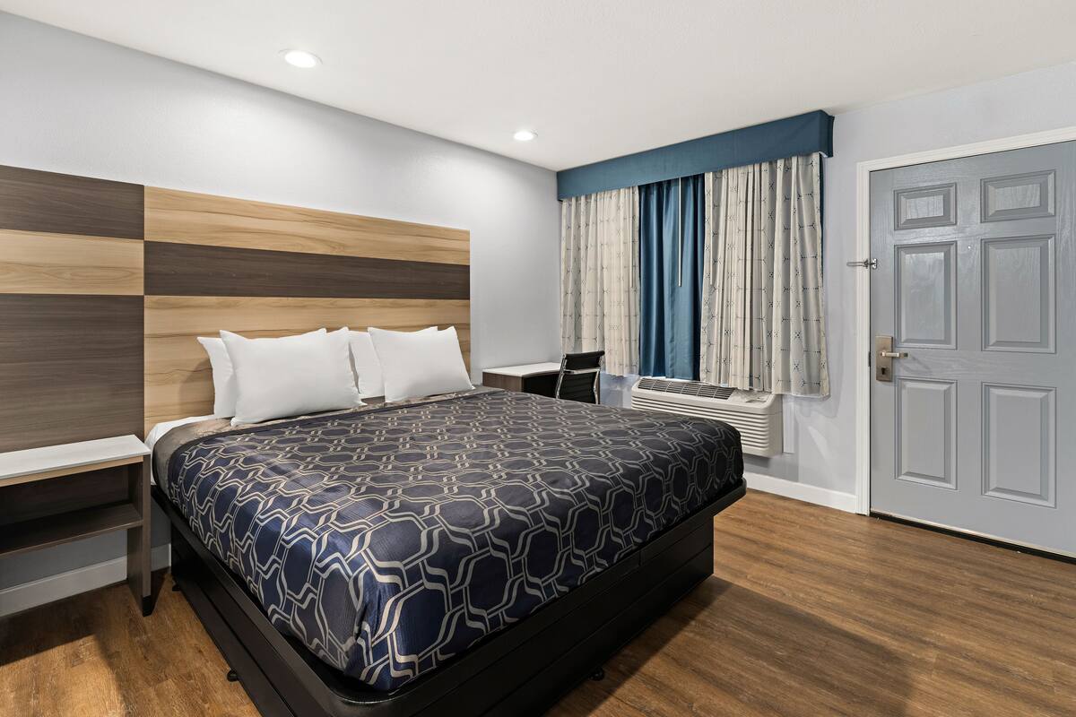 A modern hotel room with a large bed, a bedside table, a desk, and a window with curtains.