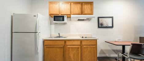 A compact kitchen with wooden cabinets, a white refrigerator, a microwave, and a small dining table with a chair.
