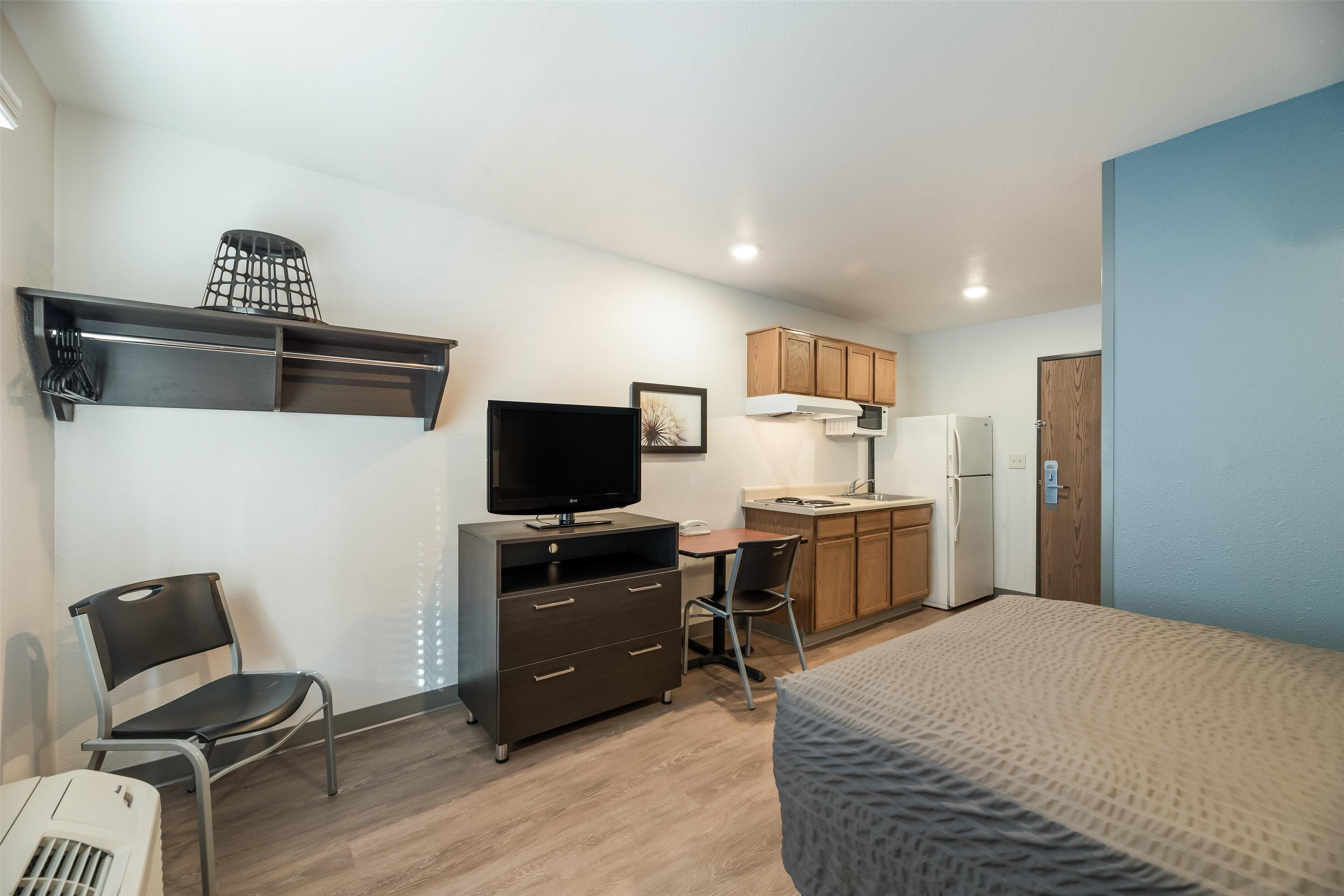 A compact hotel room with a bed, a desk with a chair, a television, a wardrobe, and a kitchenette.