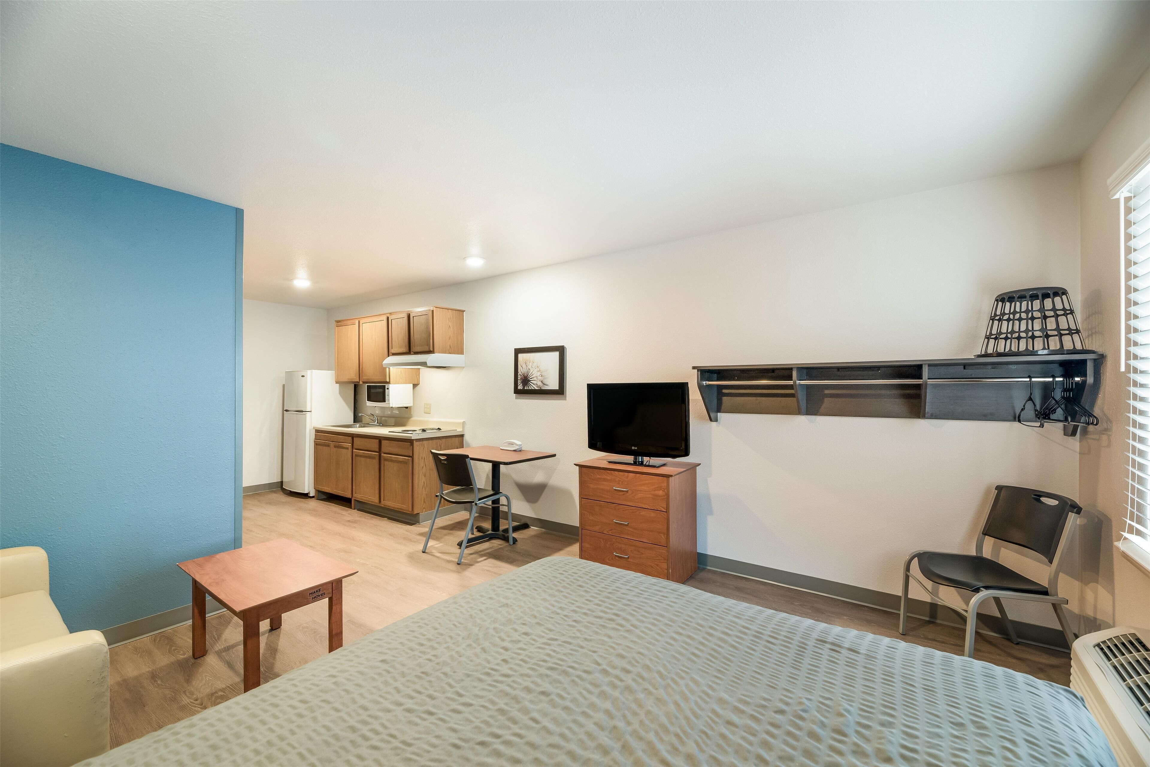 A compact hotel room with a bed, a desk, a chair, a television, a small table, and a kitchenette.