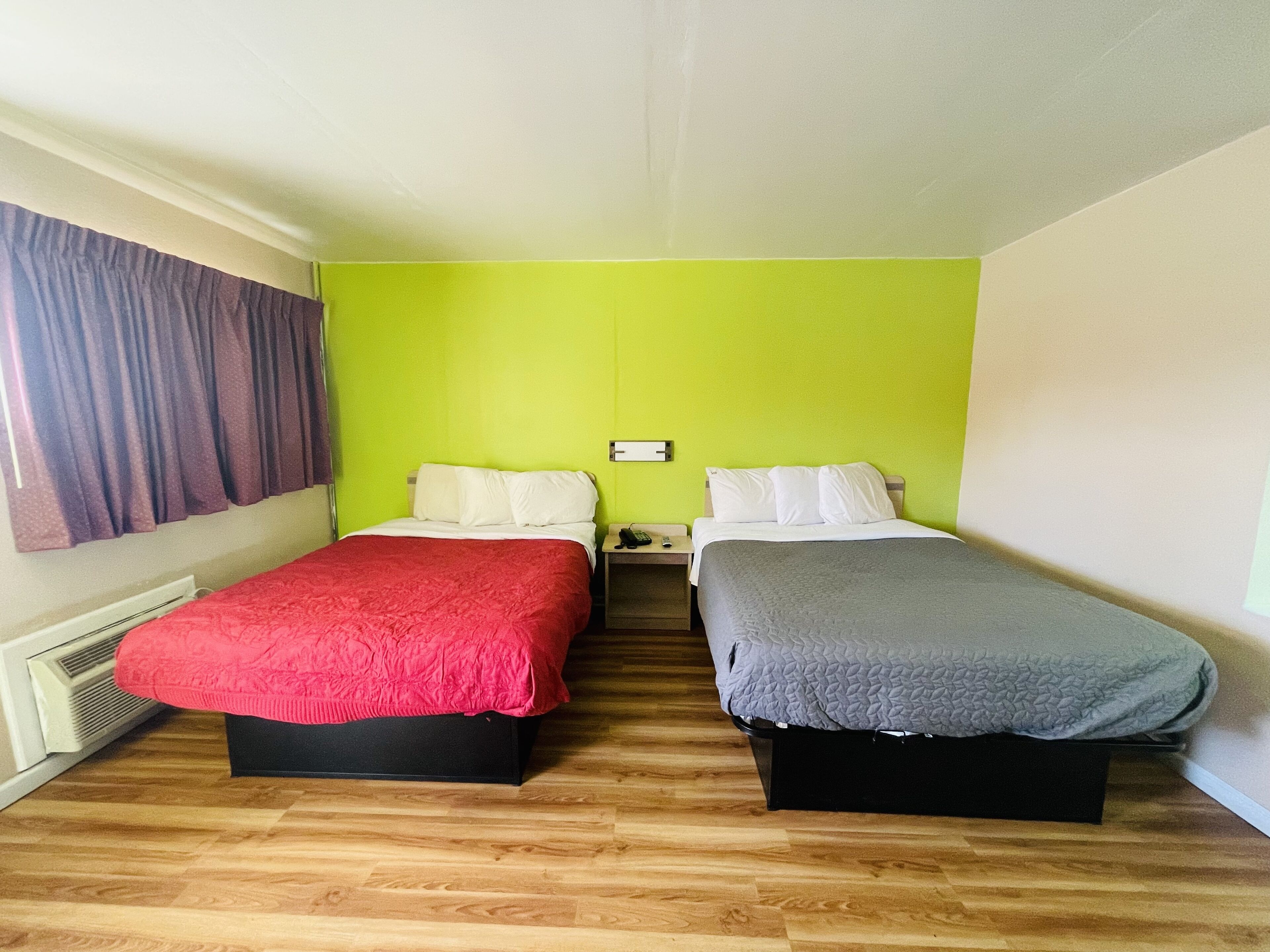 A room with two beds, each with a different colored bedspread, a nightstand with a phone and a remote, and a window with curtains.