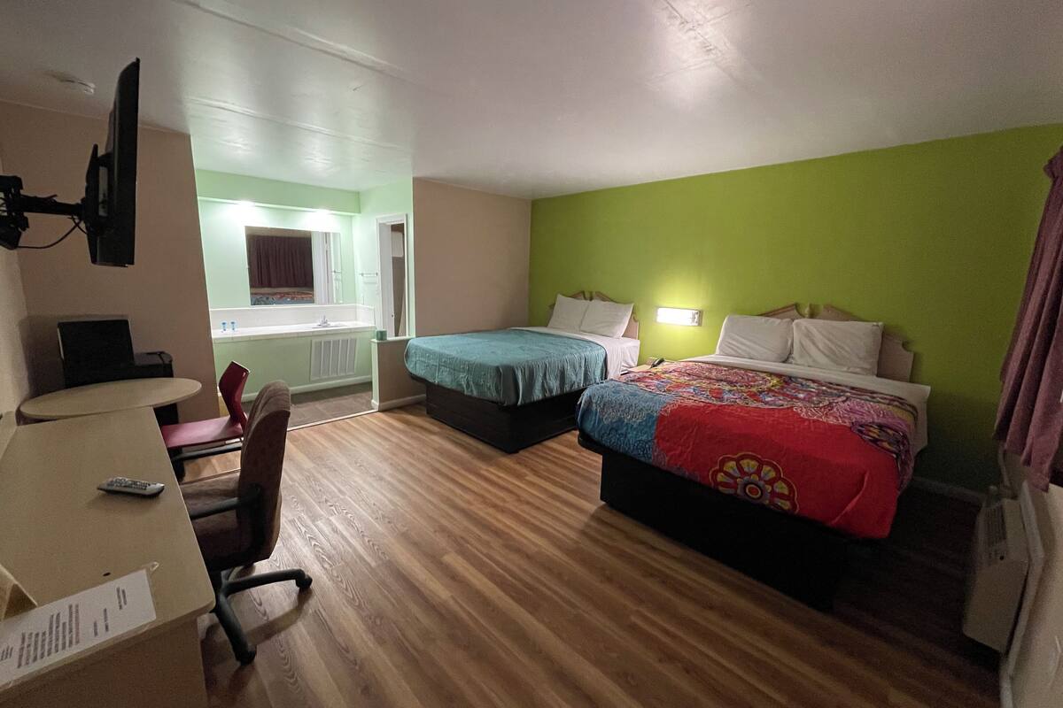 A hotel room with two beds, a desk with a chair, a small table, and a window with a view of a bathroom.