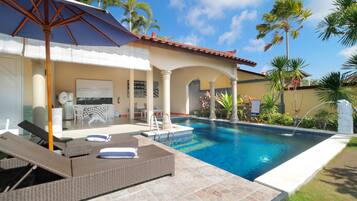 A swimming pool with lounge chairs and an umbrella, surrounded by palm trees and a tiled patio.