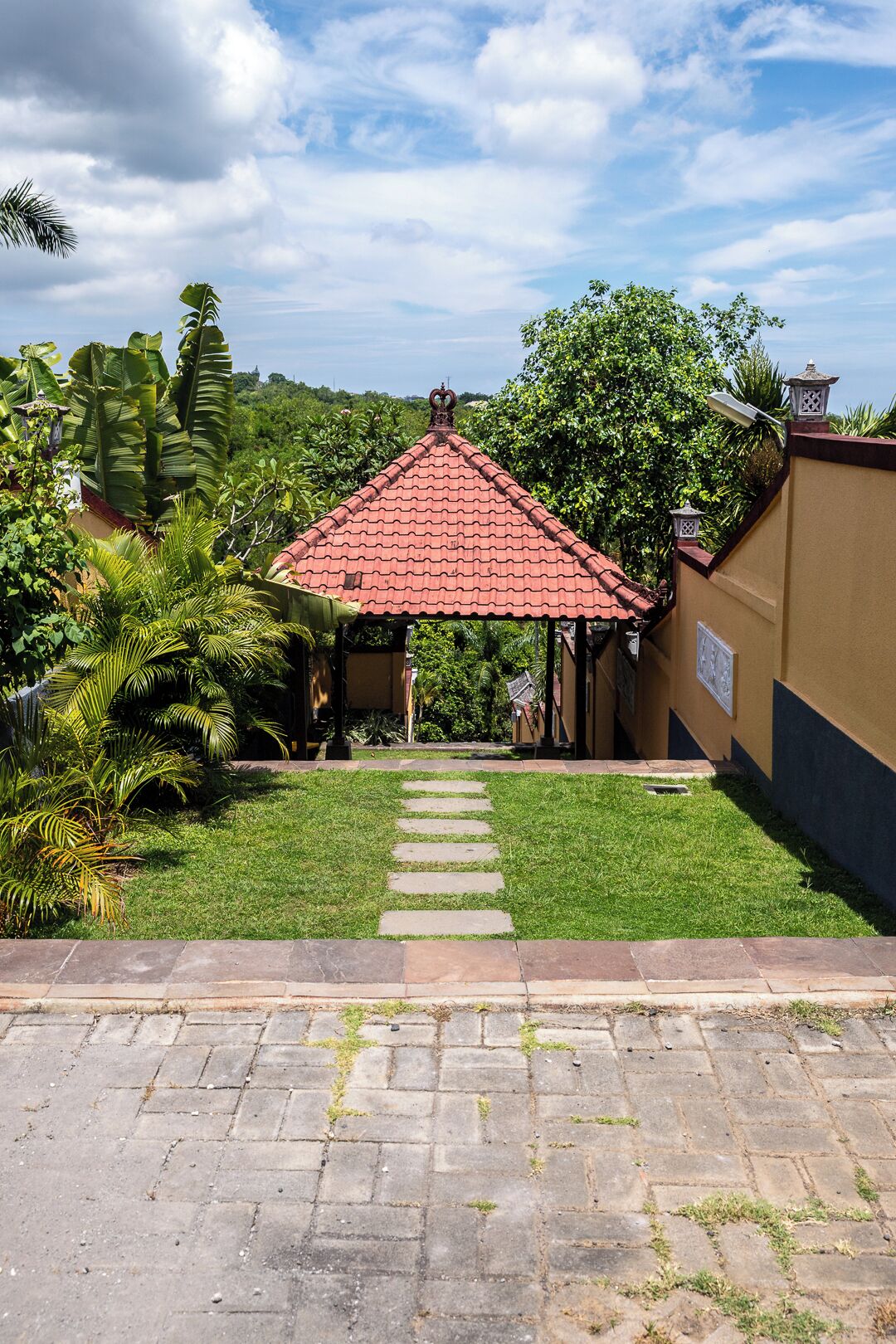 A gazebo with a red-tiled roof, surrounded by lush greenery and a paved pathway leading to it.