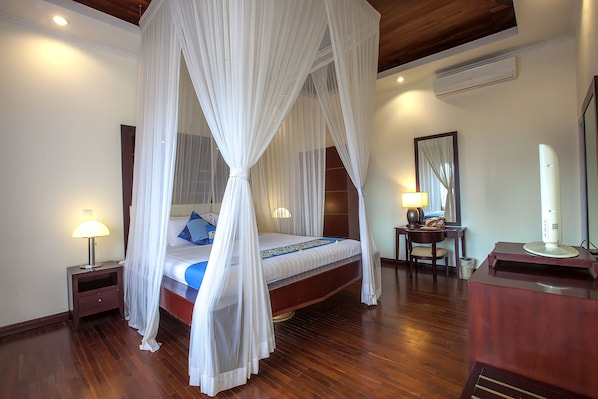 A bedroom with a four-poster bed, wooden flooring, a bedside table with a lamp, a desk with a lamp, and a mirror. - The Beverly Hills Bali a Luxury Villas & Spa (Ungasan)
