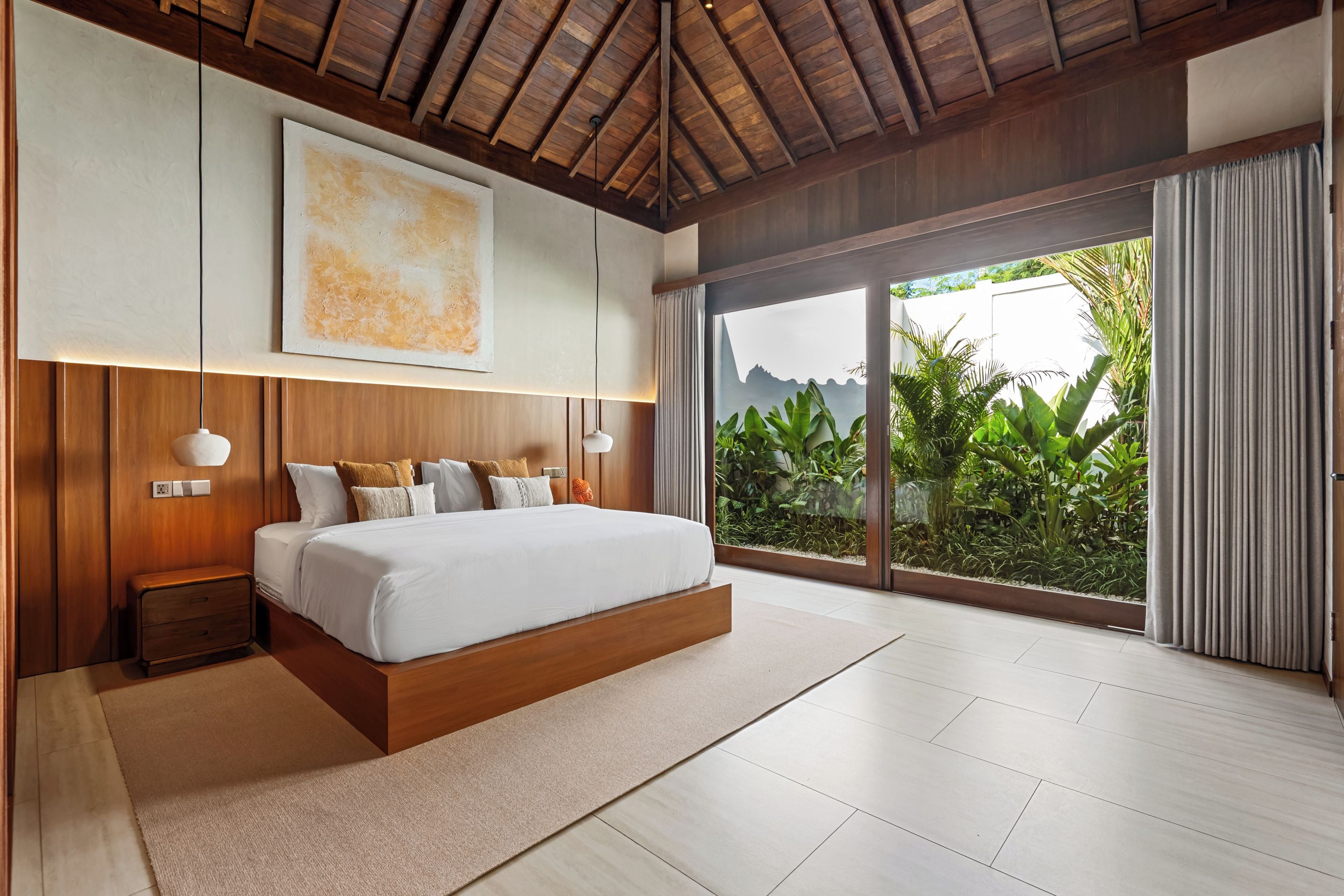 A modern bedroom with a large bed, wooden headboard, and a view of lush greenery through a sliding glass door.