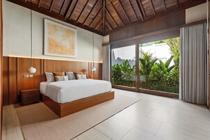 A modern bedroom with a large bed, wooden headboard, and a view of lush greenery through a sliding glass door.
