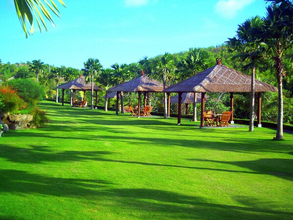 Varias gazebos con techo de paja, con asientos, rodeadas de césped verde y palmeras.