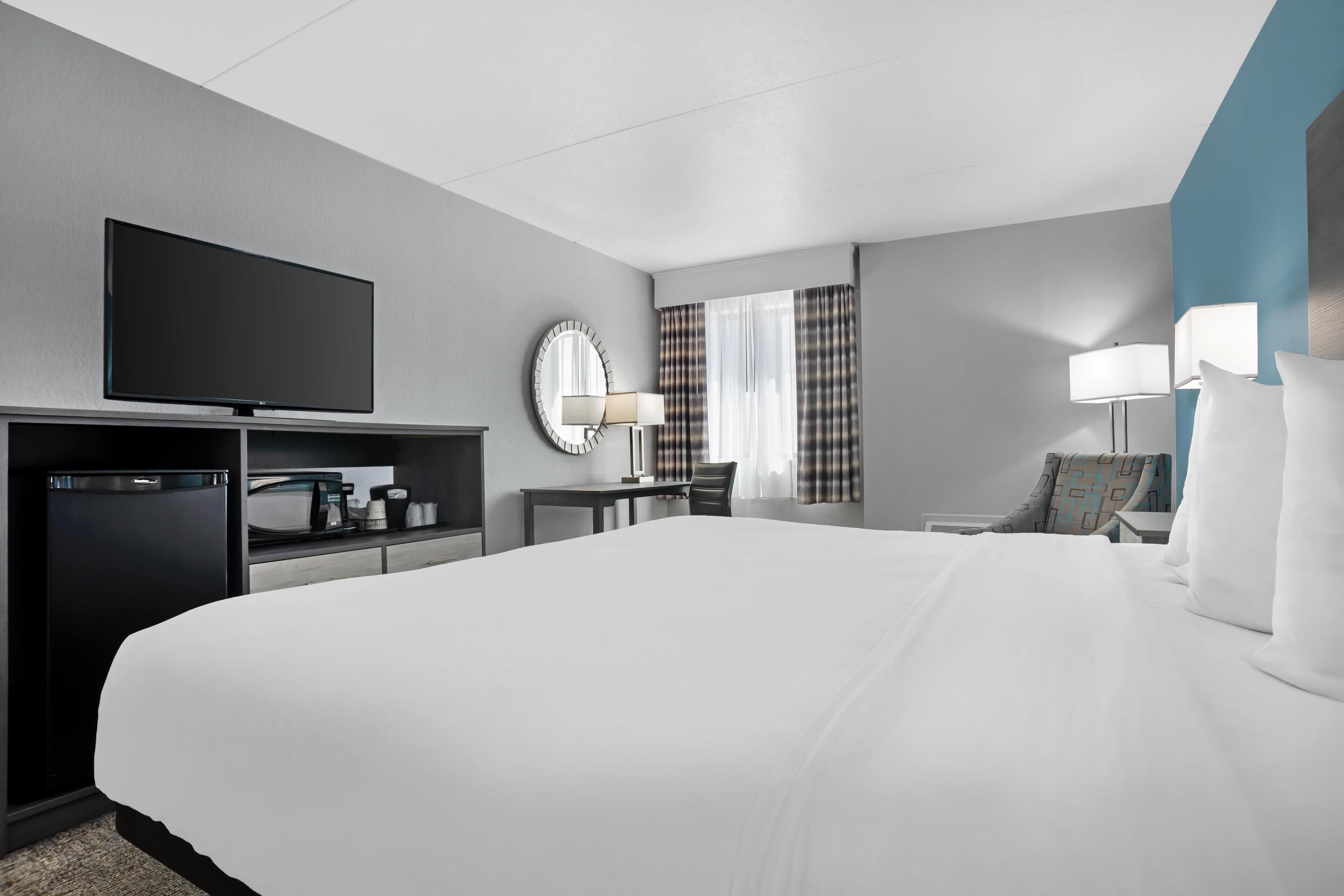 A hotel room with a large bed, a flat-screen TV, a desk with a lamp, a chair, and a small refrigerator.
