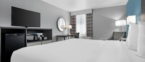 A hotel room with a large bed, a flat-screen TV, a desk with a lamp, a chair, and a small refrigerator.