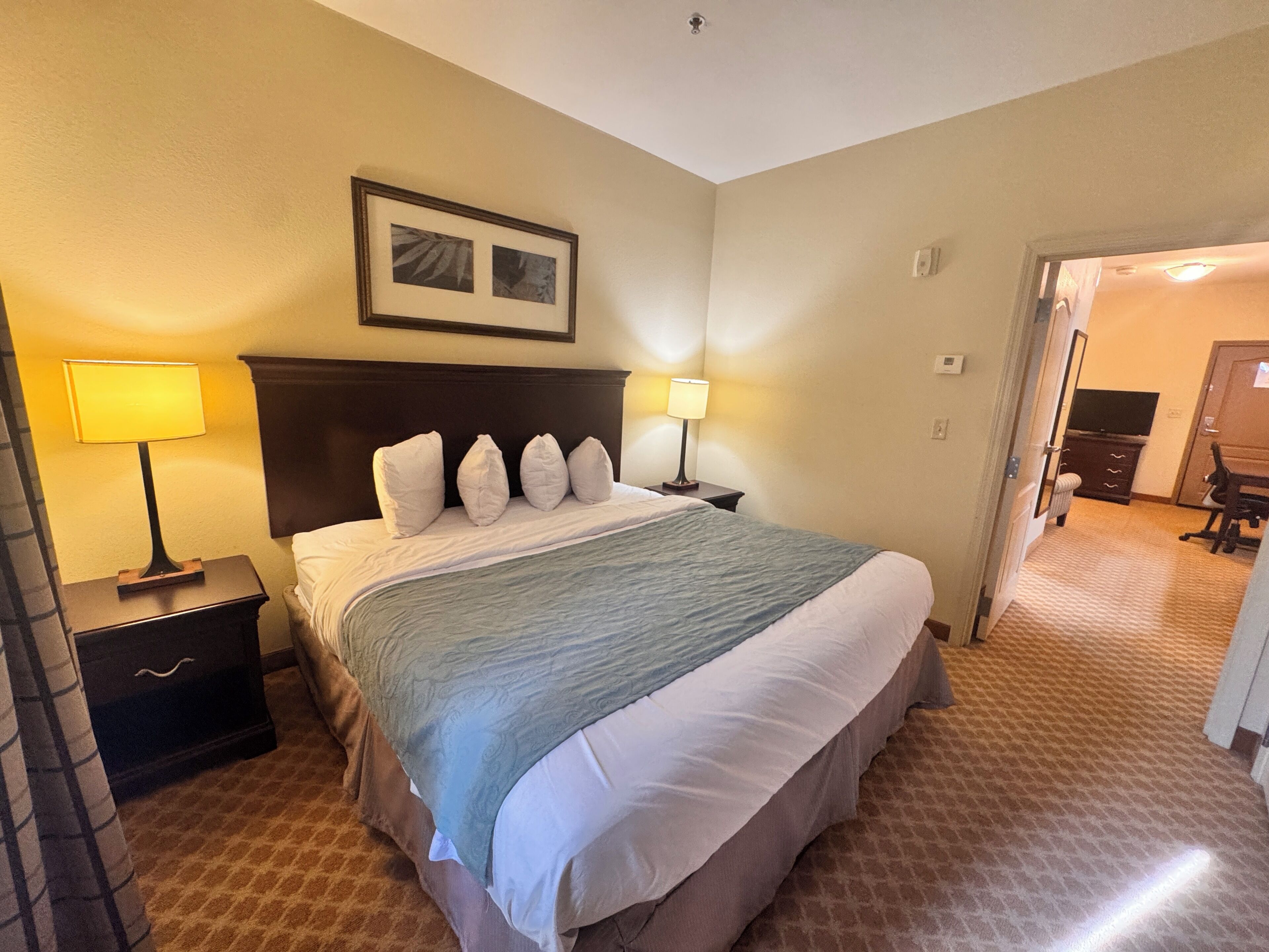 A hotel room with a bed, two bedside tables with lamps, a headboard, a framed picture above the bed, and a carpeted floor.