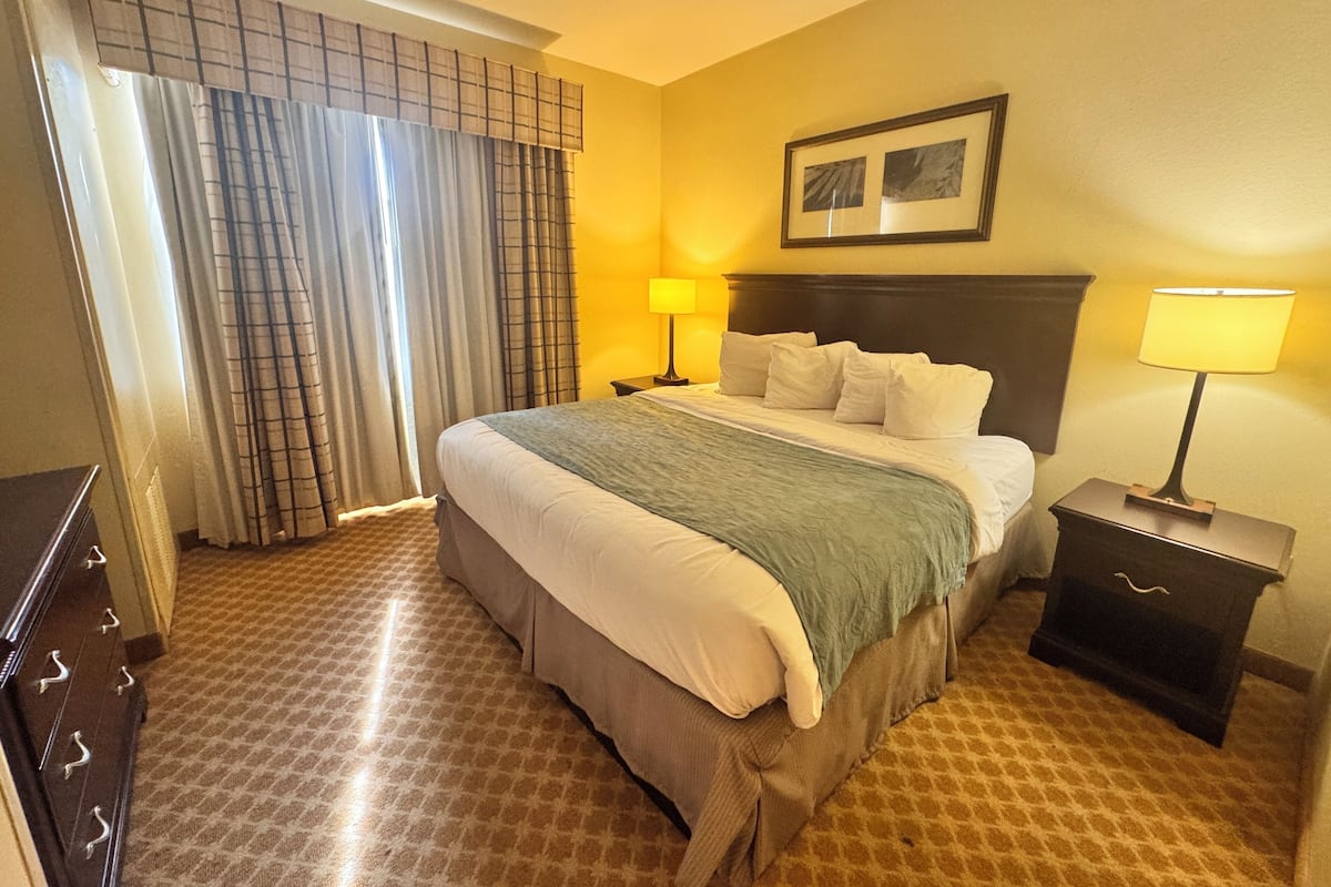 A hotel room with a large bed, two bedside tables with lamps, a headboard, a window with curtains, and a framed picture on the wall.