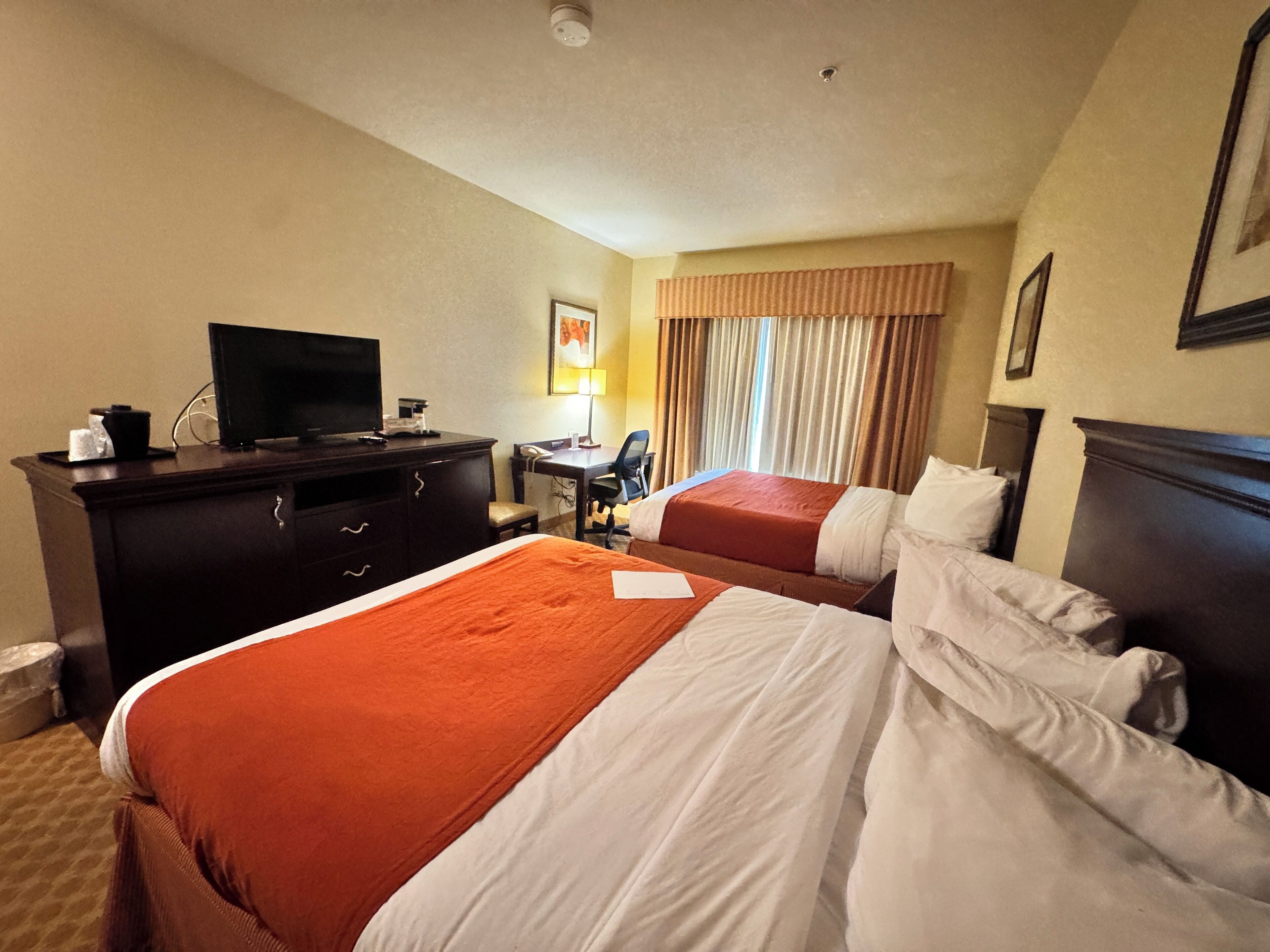 A hotel room with two beds, a desk, a television, and a window with curtains.
