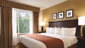 A hotel room with a large bed, a window with curtains, a nightstand with a lamp, and three framed pictures on the wall. - Hotel Pineville & Conference Center Alexandria I-49 (Pineville)