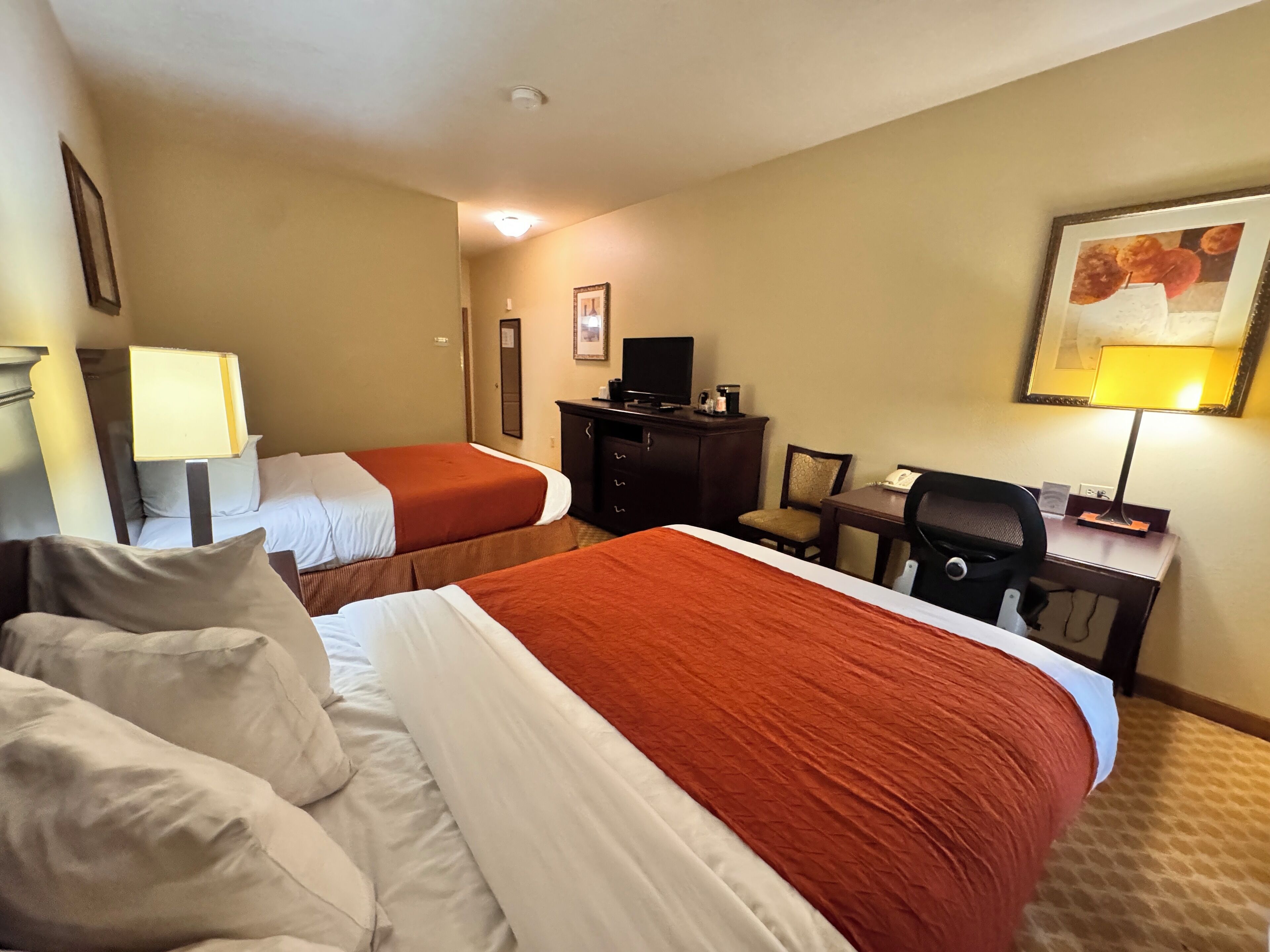 A hotel room with two beds, a desk, a chair, a television, and a lamp.