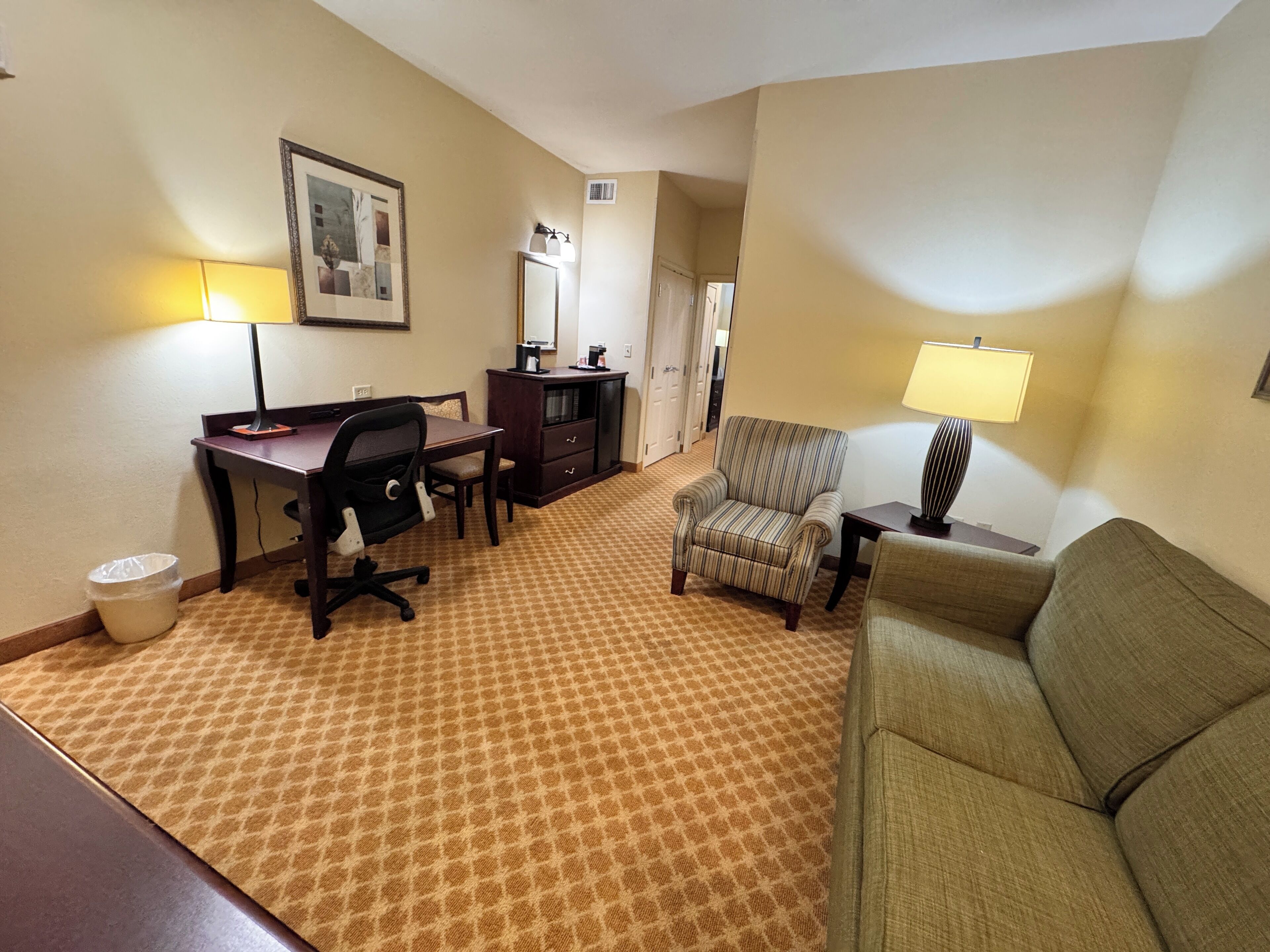 Habitación de hotel con escritorio, silla de oficina, sillón a rayas, sofá y alfombra estampada.