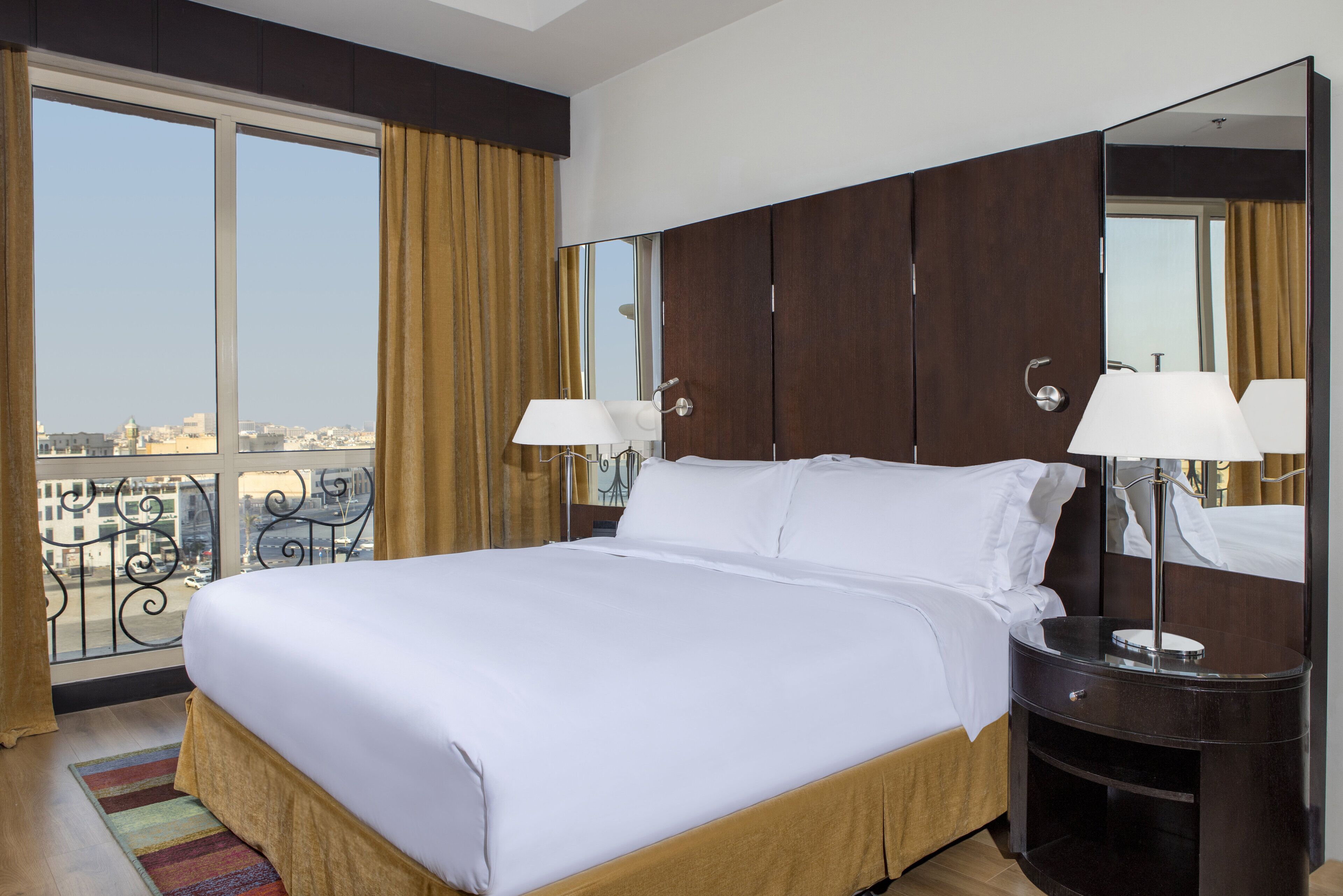 A hotel room with a large bed, two bedside lamps, a nightstand, and a large window with curtains.
