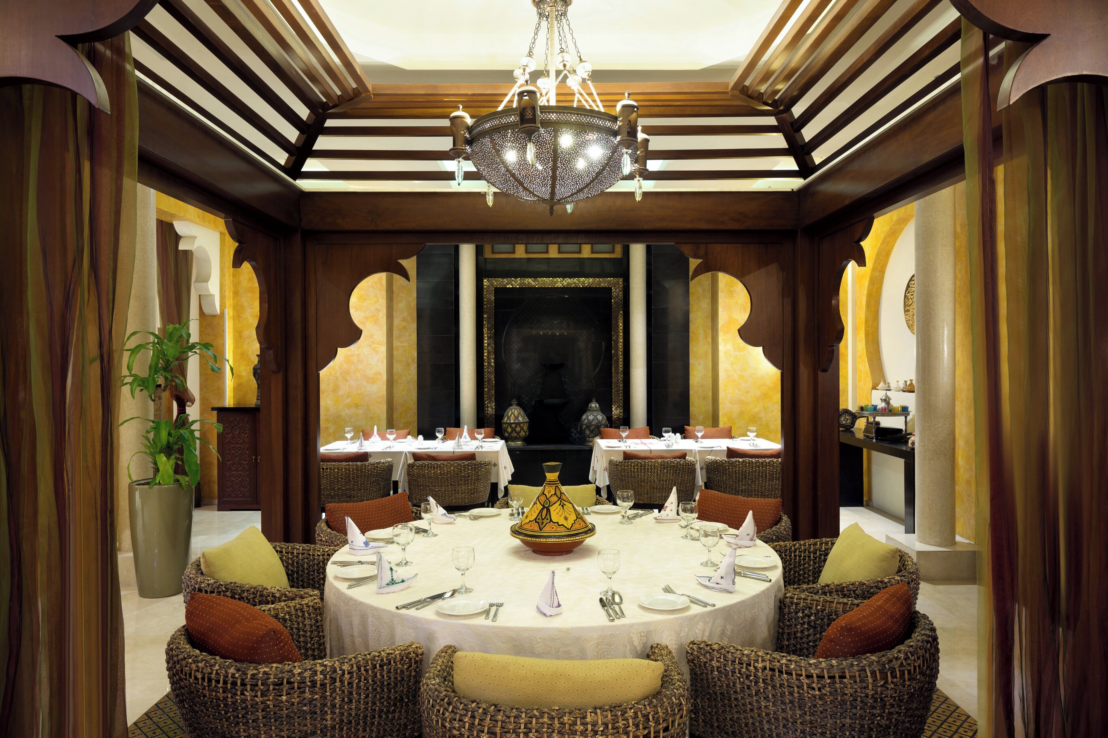 Un coin repas avec une table ronde dressée pour le repas, entourée de chaises en osier, et un grand lustre décoratif au-dessus.