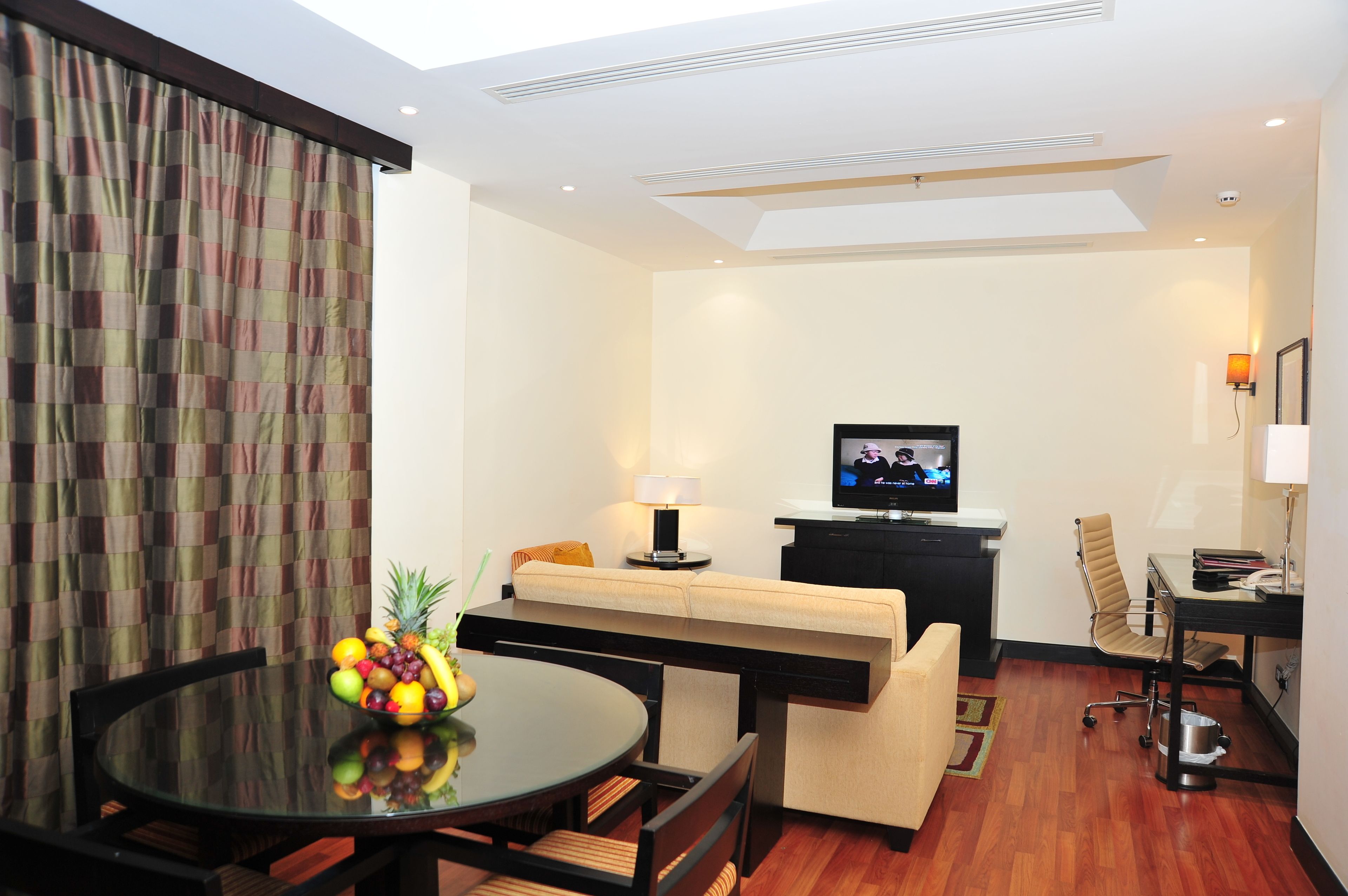 A modern hotel room with a dining area, a sofa, a TV, and a desk with a lamp.