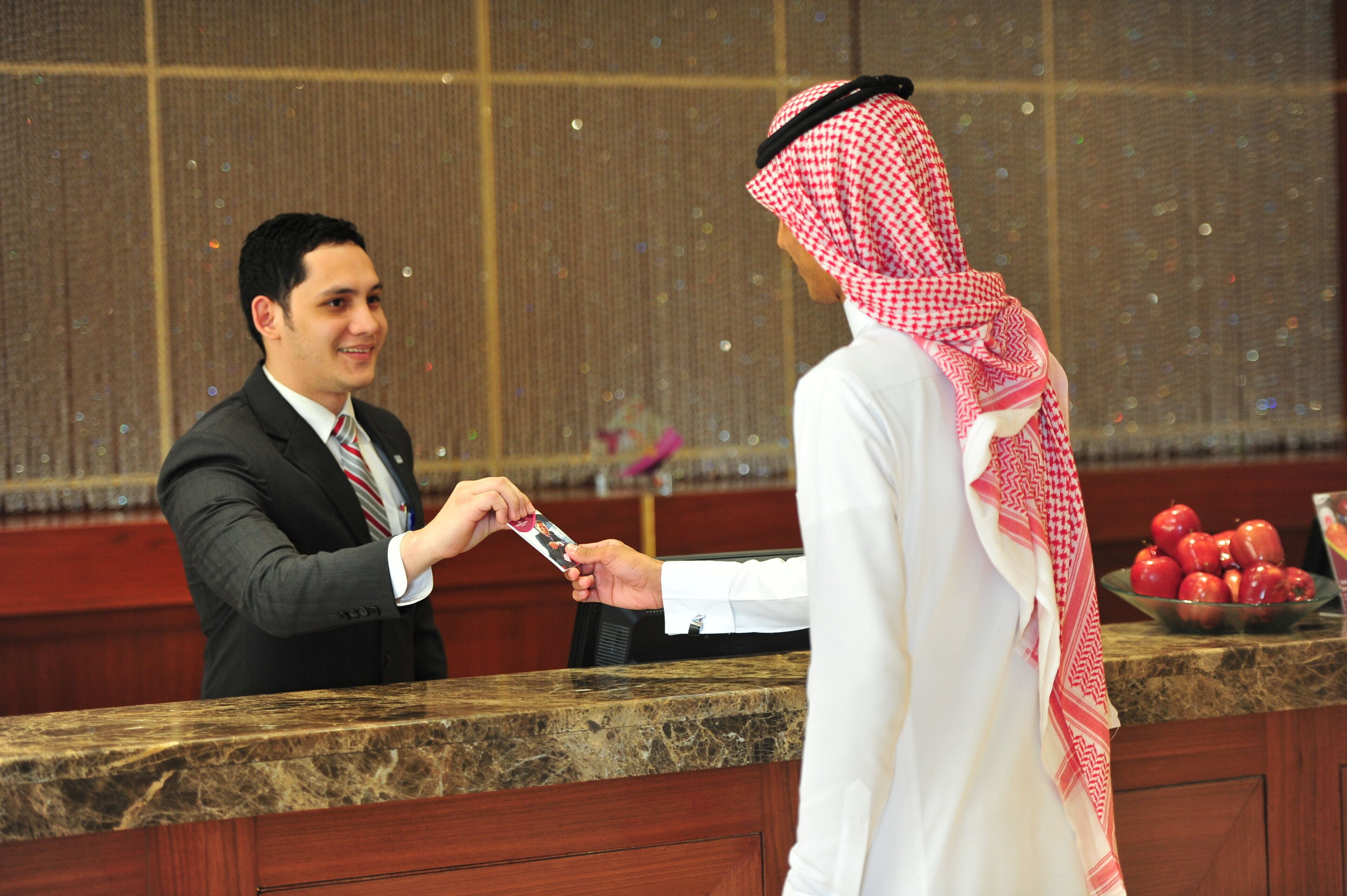 A man in a suit handing over a card to another man in traditional attire at a reception desk.