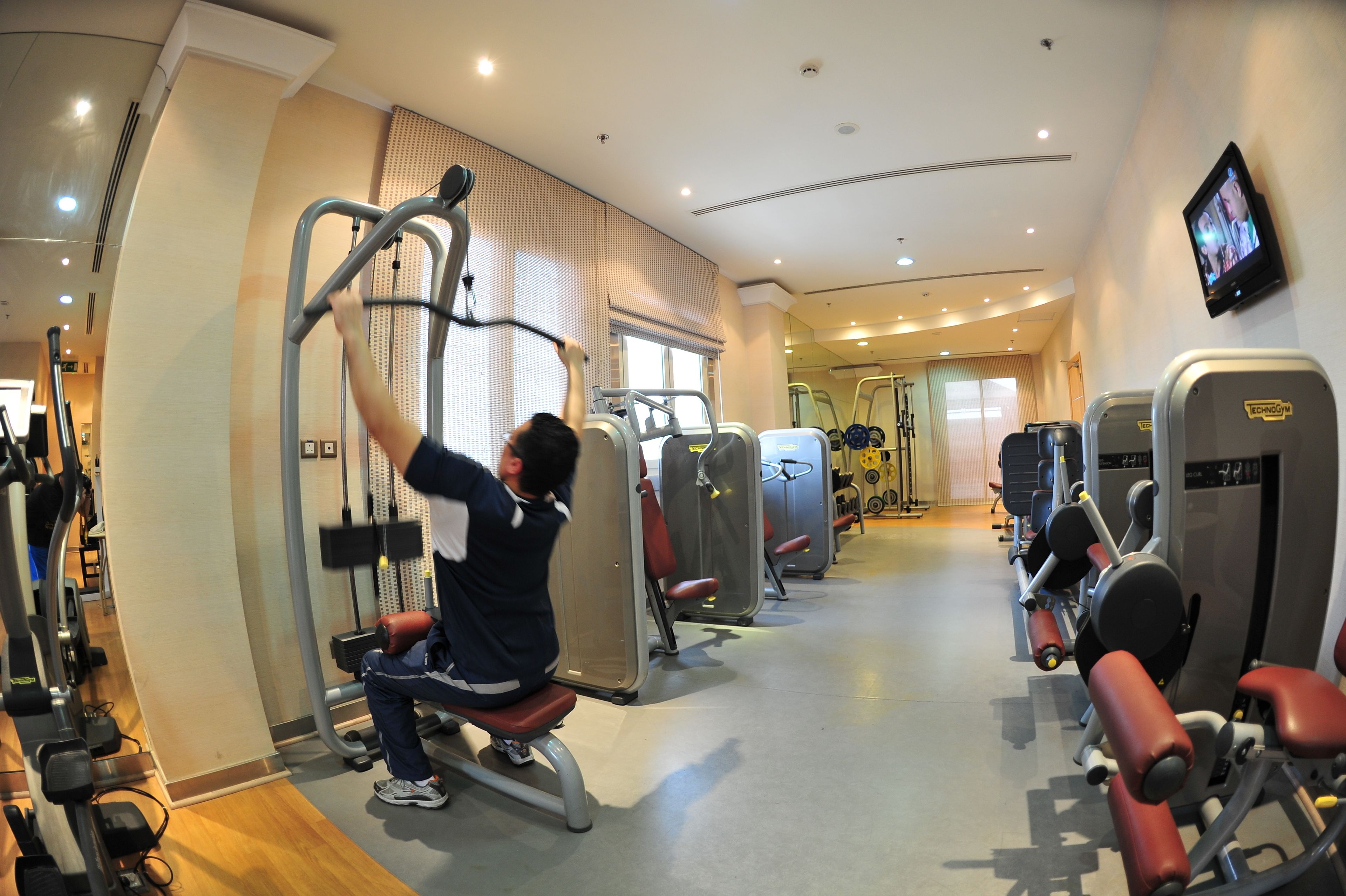 A gym with various exercise machines, a television screen, and a window with blinds.
