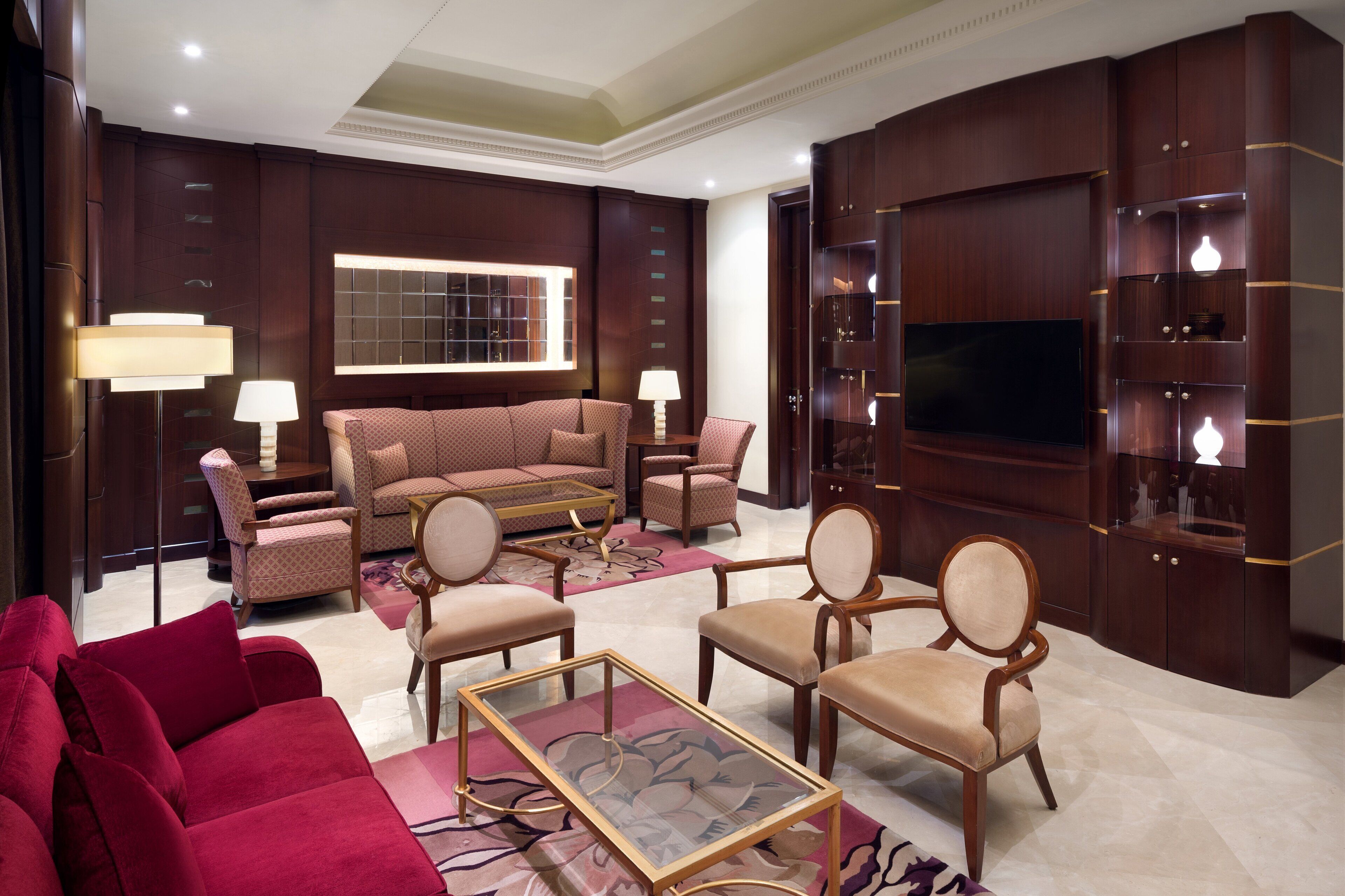A spacious living room with a large flat-screen TV, comfortable seating including a red sofa and beige armchairs, and a wooden entertainment unit with shelves.