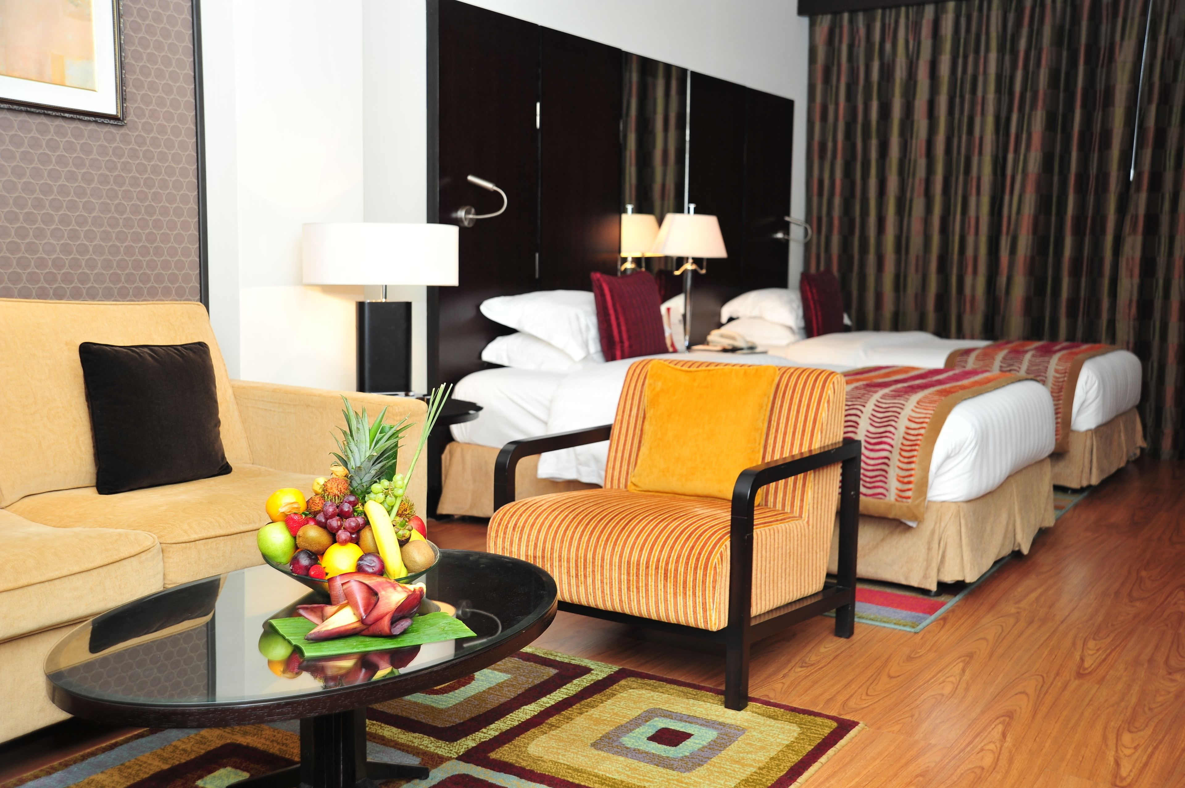 Une chambre d’hôtel avec un canapé, un fauteuil, un lit avec deux oreillers, une table de chevet avec un panier de fruits et une lampe.