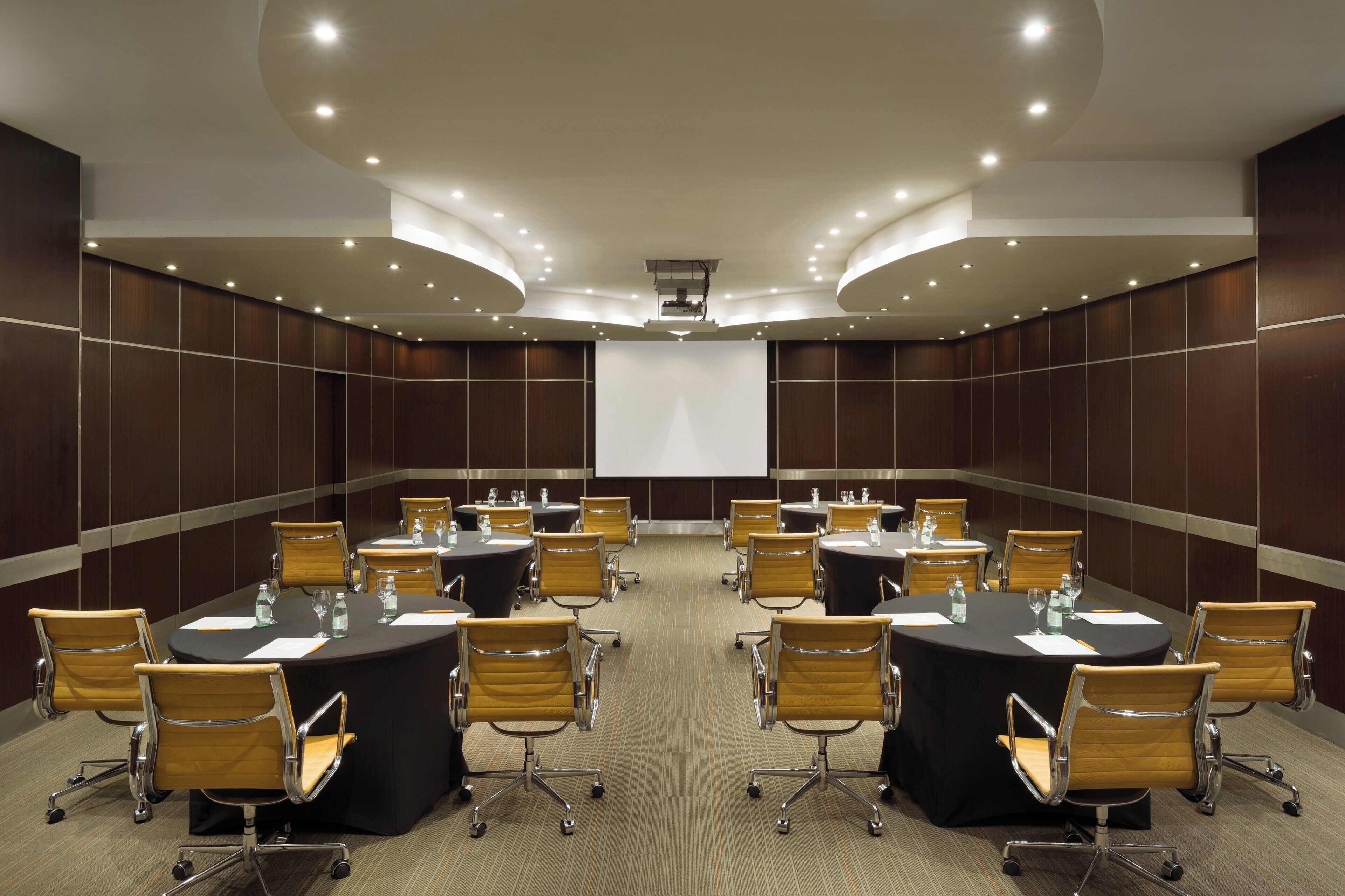 A conference room with multiple tables and chairs, equipped with water bottles and glasses, and a large screen at the front.