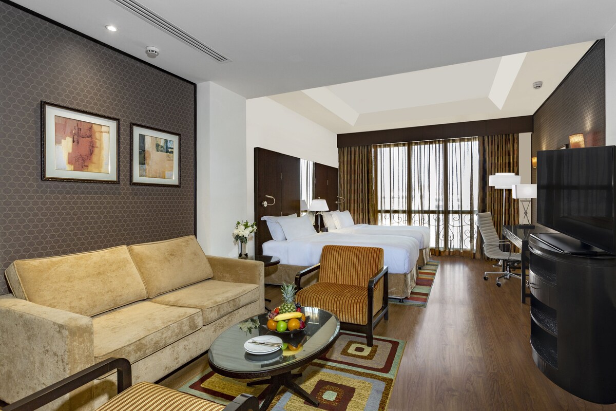 A hotel room with a sofa, a dining table with fruit, a bed, a TV, and a large window with curtains.
