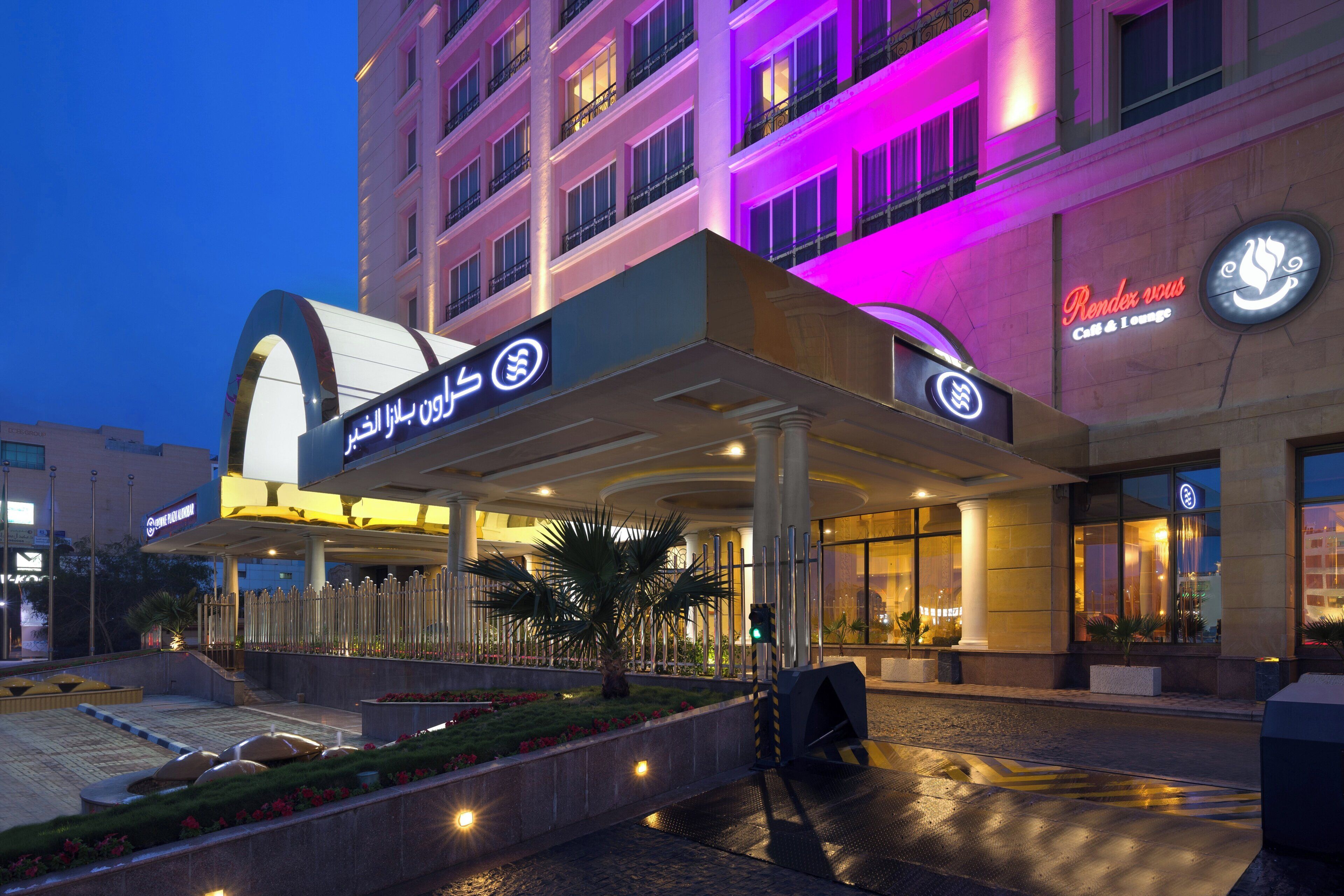 A hotel entrance with a covered walkway, palm trees, and a sign for 'Rendezvous Café & Lounge'.