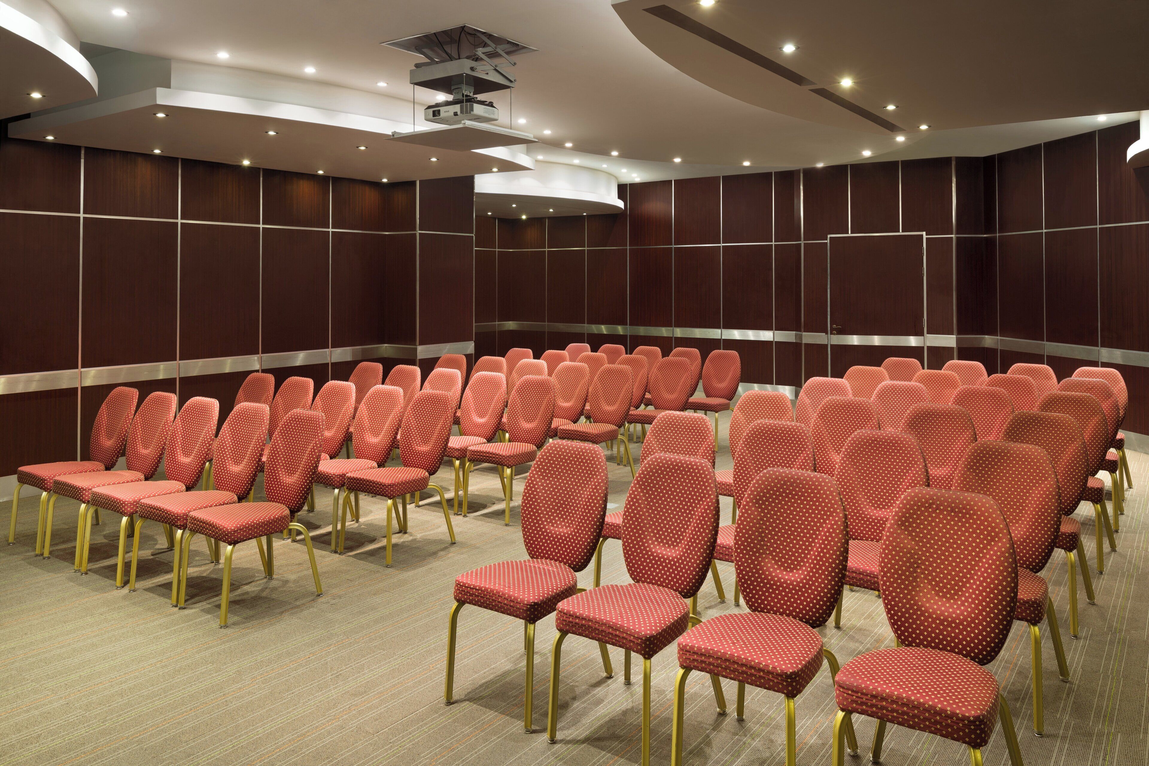 A conference room with rows of chairs, a ceiling projector, and recessed lighting.